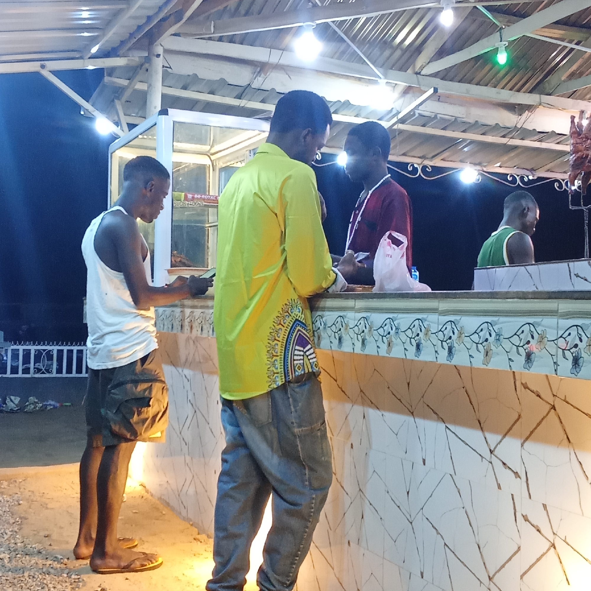 Four dark-skinned men are standing at a counter at a food stand in Akwanga, Nigeria. The man on the left is wearing a white tank top and khaki shorts with yellow flip-flops, looking at a phone screen. A man in a bright yellow patterned shirt faces forward. Another man is in a maroon shirt. The last man is in a green tank top. The counter has a floral pattern and is tiled with a marble-like crack pattern. Above the counter is a glass display case with what appears to be food inside. The roof of the stand is made of metal sheets and has exposed wooden beams with light bulbs. Hanging to the right is meat. The ground is dirt and gravel. There is a white fence visible in the background.