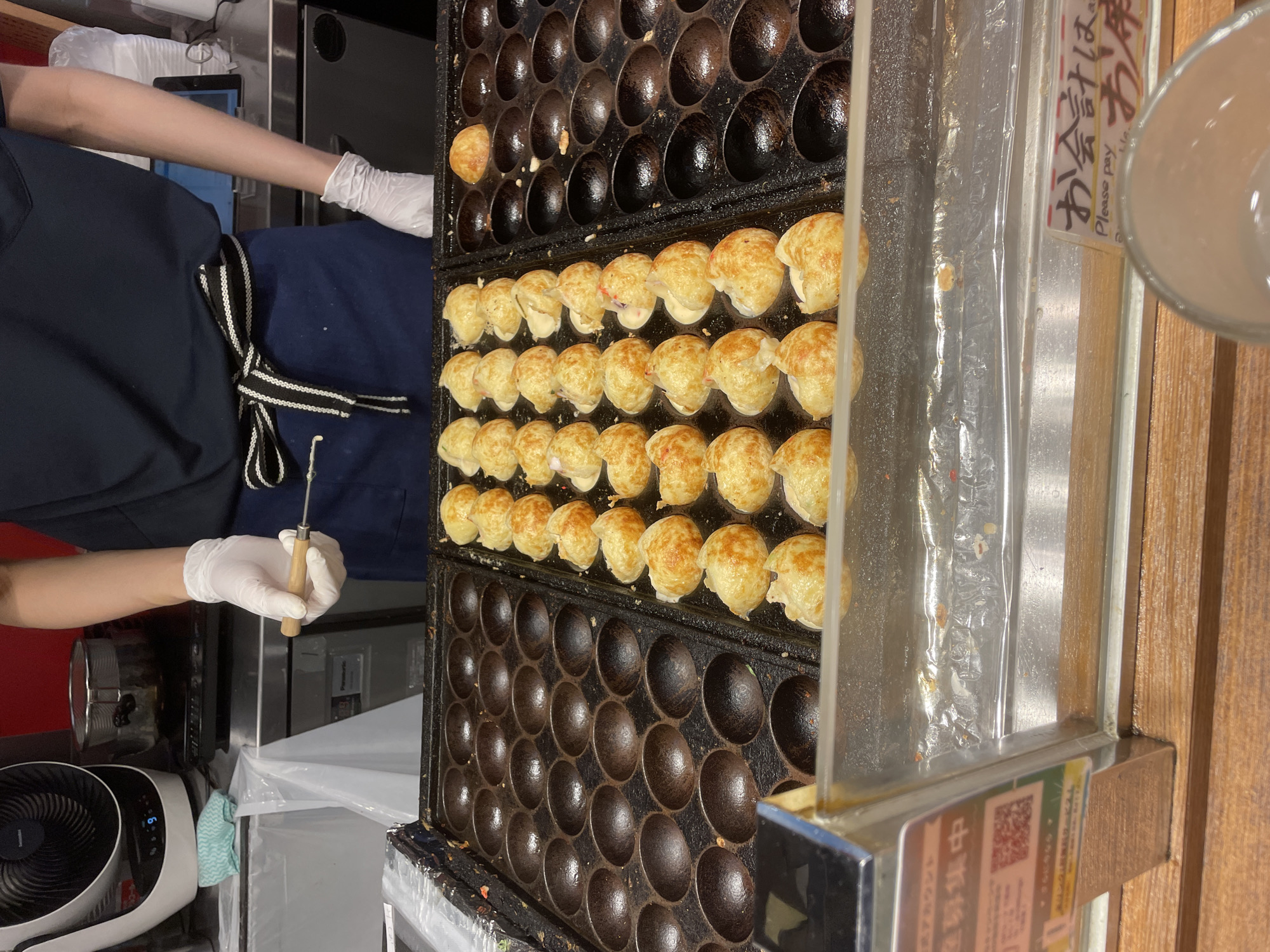 A person wearing white gloves and a dark blue apron with a striped sash is cooking takoyaki in a specialized pan at a food stall in Taito, Japan. The person is holding a skewer with a wooden handle, presumably to turn the takoyaki. In the foreground, rows of half-cooked takoyaki balls are visible in the indented pan. To the left of the takoyaki pan, there are empty indentations in another section of the pan. Behind the person, a white fan and a stainless steel pot are visible on a counter. A sign with Japanese characters and a QR code is attached to a clear plastic barrier in front of the stall. Another sign with Japanese text and "Please pay" is also visible on the barrier. The overall environment appears to be a food preparation area in a public space, likely a market or street food vendor.