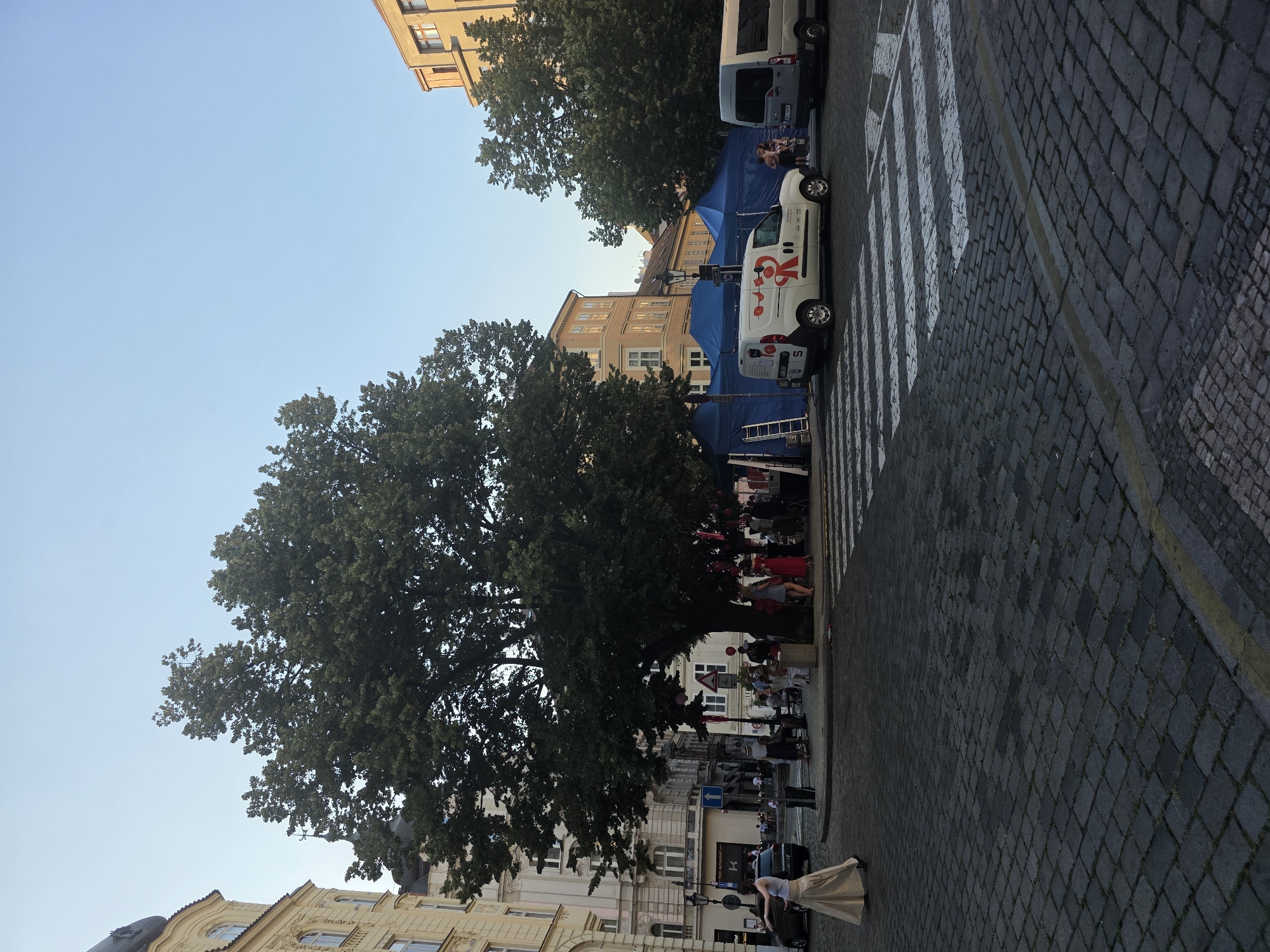 The image depicts a street scene, likely in a European city, during daylight hours. The sky is clear and blue, suggesting fair weather. A large, leafy tree dominates the left side of the frame, casting shadows onto the cobblestone street and adjacent buildings. Several people are gathered under the tree, and some are seated at tables, indicating a social event or gathering.
Two white vans are parked on the right side of the street, one closer to the foreground. The van in the foreground has a red and white logo with stylized figures on its side, along with some visible text, though it's difficult to read clearly. A ladder leans against the blue tent behind the vans, suggesting some sort of setup or work is in progress. The street features a prominent pedestrian crossing with white stripes.
The buildings lining the street are multi-story and appear to be of historic architecture, with various window styles. The overall atmosphere seems relaxed and casual, with people enjoying themselves outdoors. The time of day appears to be late afternoon or early evening, given the angle of the sunlight.