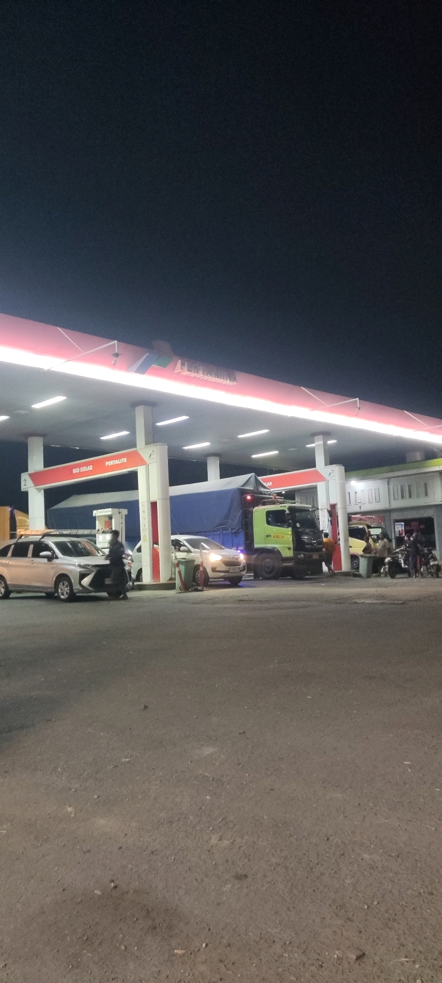 A nighttime exterior shot captures a Pertamina gas station in Pelayangan, Indonesia. A red and white awning with the Pertamina logo stretches across the frame, illuminated by fluorescent lights. Beneath the awning, several vehicles are present.
On the left, a silver car with its driver's side door open is parked near a gas pump. A person in dark clothing is standing next to the car, appearing to interact with it. Further down, a white car is parked at another pump, its headlights on.
A large green and black truck with a blue tarp-covered cargo bed is positioned centrally, also at a gas pump. Several people are visible around the truck, some appearing to be workers. To the right of the truck, a yellow car is parked, and a group of people are gathered near a building that appears to be a convenience store or office associated with the gas station. Motorcycles are also present on the right side of the scene. The ground in front of the station is paved asphalt. The sky above is dark.