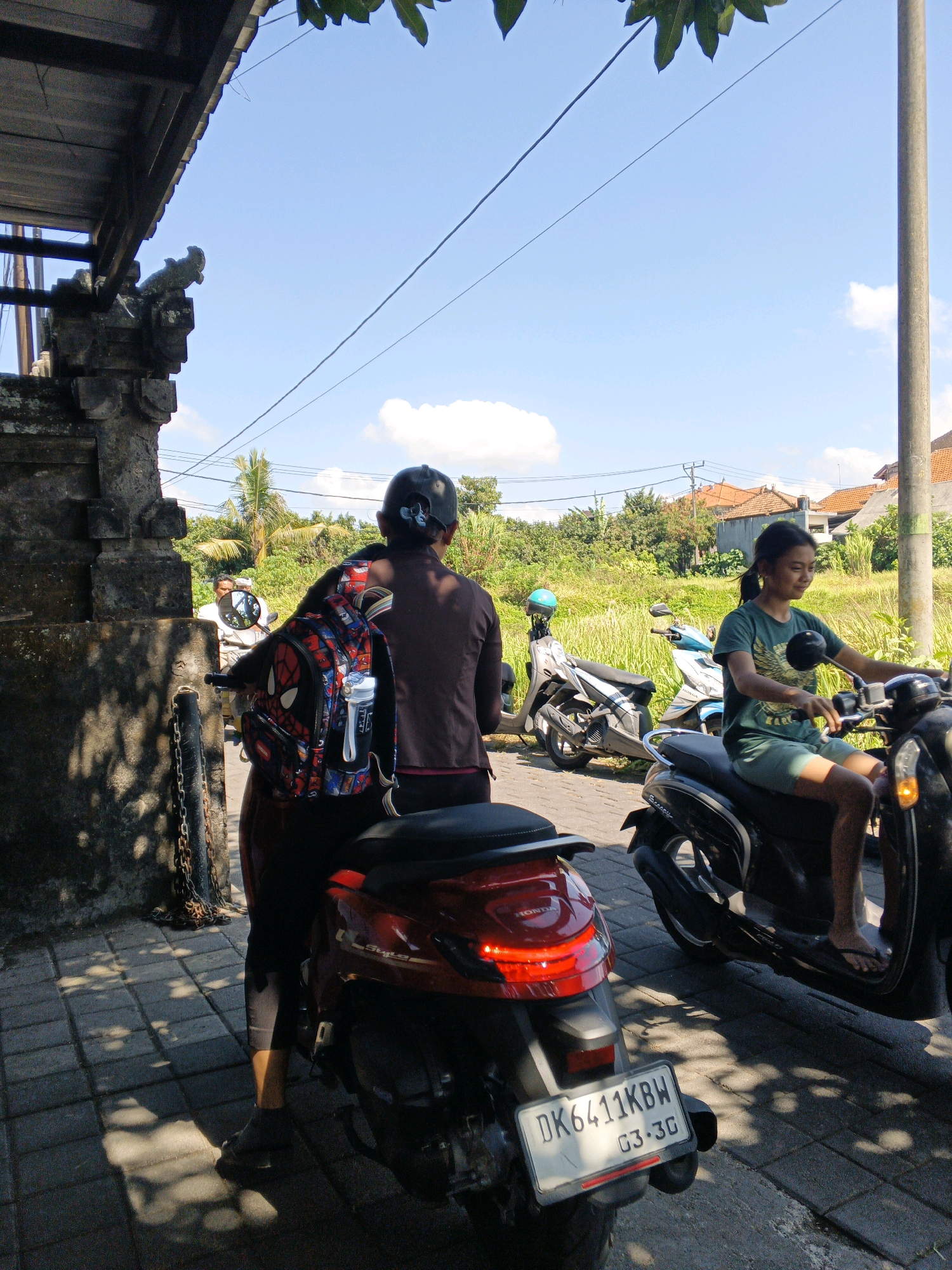 In Sukawati, Indonesia, under a clear blue sky dotted with white clouds, two individuals are observed near a traditional stone structure. A person wearing a dark shirt and a black cap is seated on a red scooter, their back to the camera. A backpack, adorned with a Spider-Man design, is attached to the scooter's rear. To the right, a young woman with dark hair tied in a ponytail is riding a black scooter. She wears a green t-shirt and shorts and smiles as she looks forward. Several other scooters are parked nearby amidst lush green vegetation and buildings with terracotta roofs. The ground is paved with stone tiles, and shadows from overhead foliage create a dappled pattern. A pole and power lines are visible in the background, indicating an urban or semi-urban setting. The license plate on the red scooter reads "DK 6411 KBW 03-3G."
