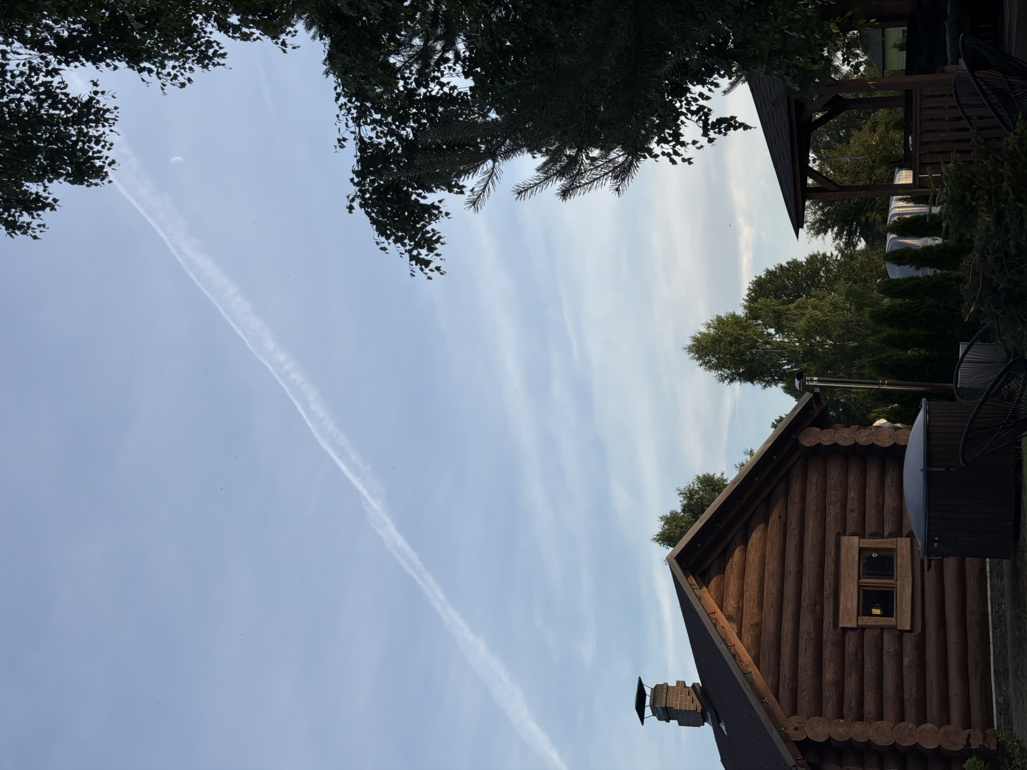 The image shows a log cabin in Dreiliņi, Latvia, with a hot tub in the foreground. The cabin has a dark roof and a brick chimney. A trail of white clouds stretches across the light blue sky, with a crescent moon visible. Lush green trees frame the scene. In the background, there is a covered gazebo and other structures, suggesting a residential or recreational area. The overall impression is one of a peaceful, rural setting.