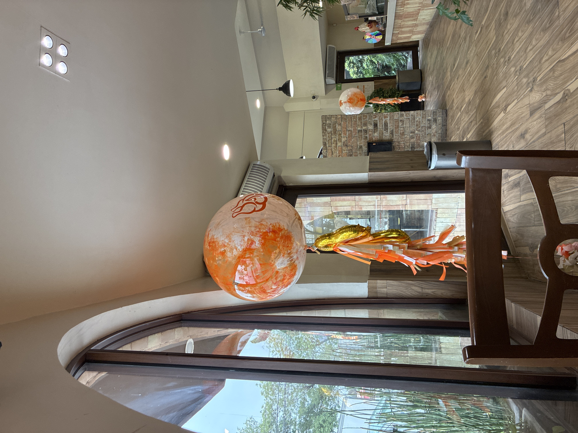 The image shows the interior of a building, possibly a restaurant or cafe, in Cuautitlán Izcalli, Mexico. The camera angle is rotated, giving a slightly disorienting perspective. A large, round, orange and white balloon with decorative streamers is suspended near a large arched window. Through the window, trees and power lines are visible, suggesting an urban or suburban environment. The interior has wooden floors and brick walls. In the background, there is a window that looks out onto a green scene. Part of a wooden chair is in the foreground. Ceiling lights and other hanging decorations are also visible in the background.
