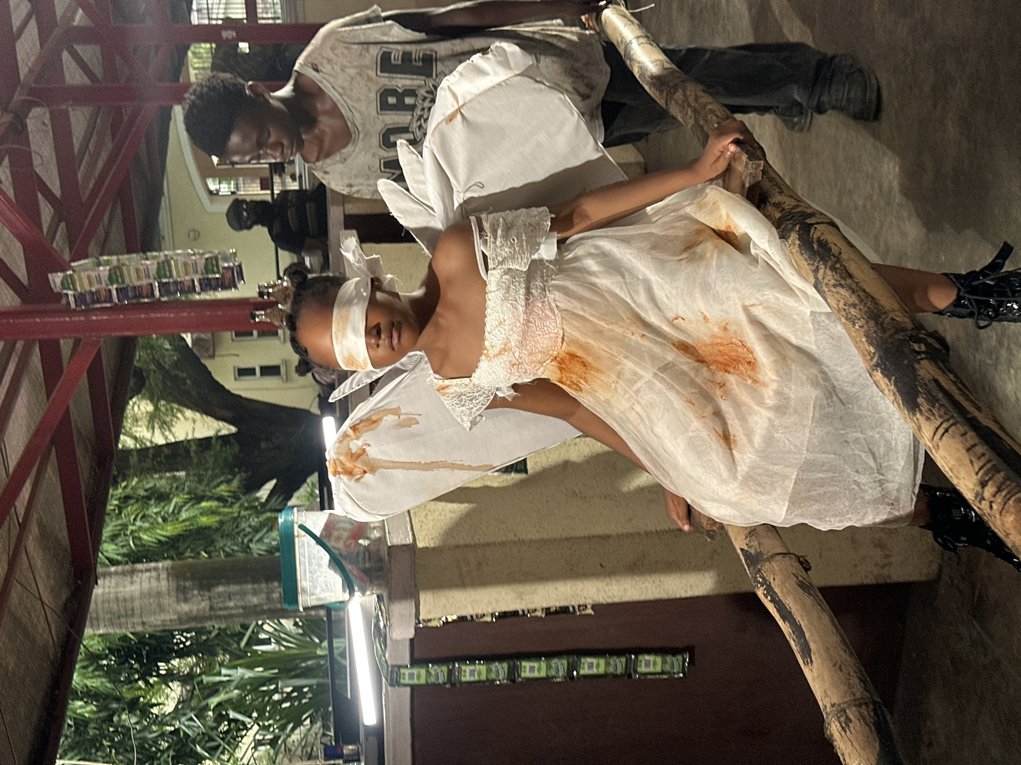 A young, dark-skinned girl, blindfolded, wearing a soiled white dress and rudimentary white wings, is positioned on a rough-hewn bamboo structure. Orange stains mar the front of the dress and wings. She is supported by a dark-skinned male wearing a grey t-shirt and dark pants. The location appears to be an outdoor space possibly in Lagos, Nigeria, with glimpses of a red-roofed structure, greenery, and an adjacent building with lighted windows. Items resembling packaged goods are affixed to a nearby wall.