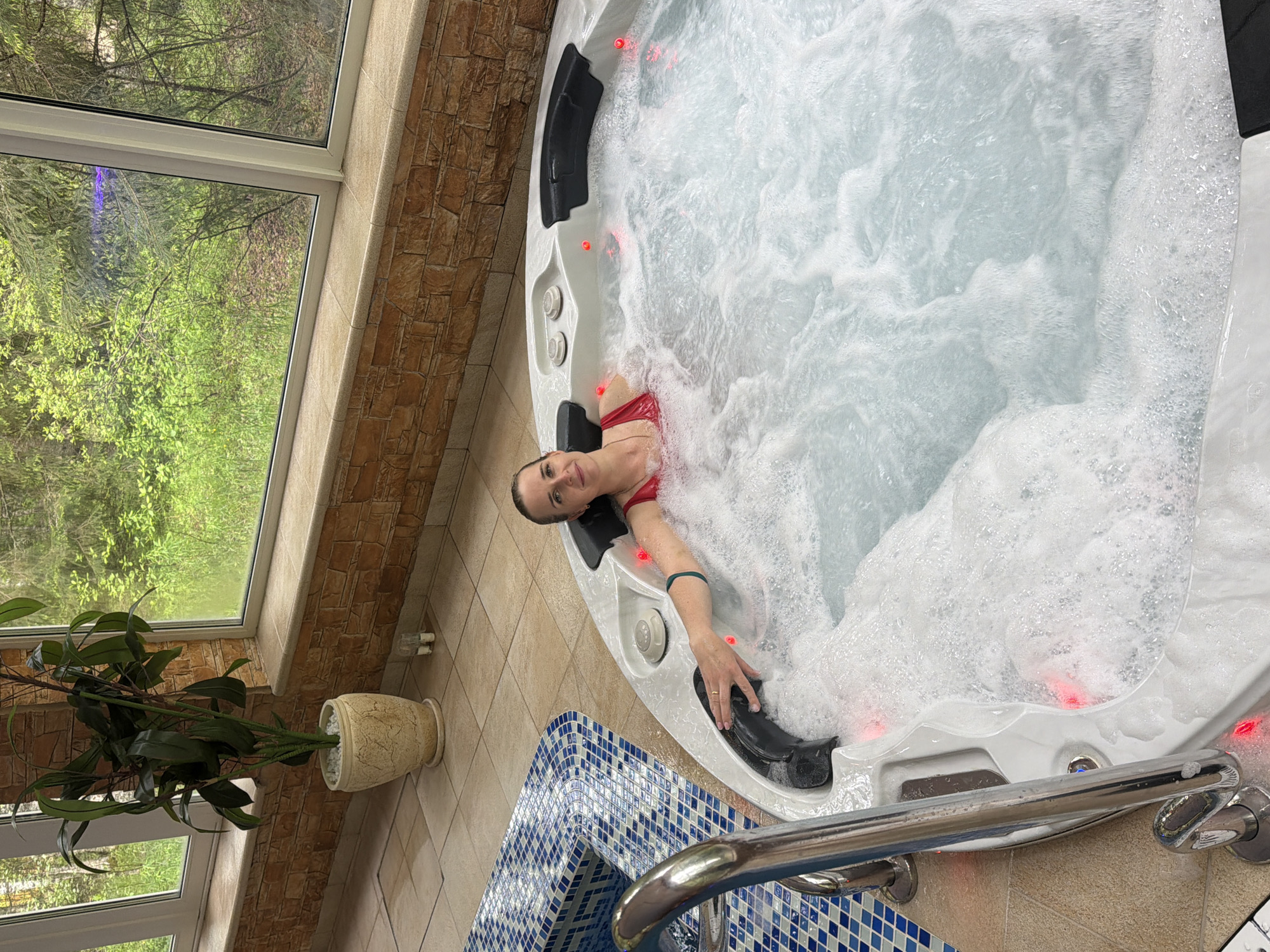 A woman is in a white hot tub, partially submerged in bubbly water. She wears a red swimsuit and a black headrest. Her arm rests on a black fixture within the tub. The hot tub is located indoors with a window showing a view of trees outside. The tub sits on a tiled floor, bordered by a blue and white tiled structure with a silver railing. A potted plant stands near the window. The walls near the window are covered in a stone-like material. The scene is indoors, likely in a spa or wellness center, potentially located in Zaplav'e, Russia, as per the user's request.