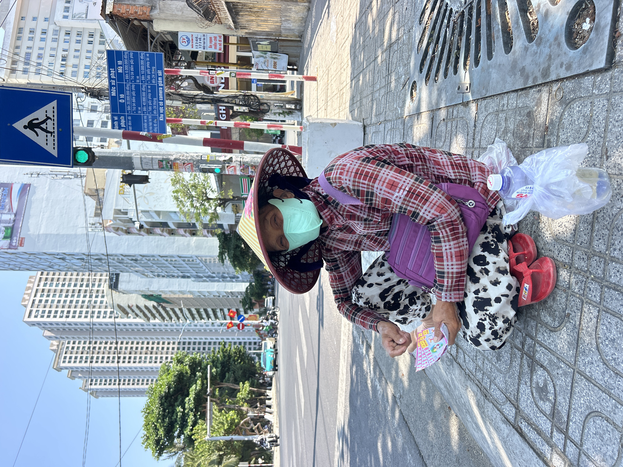 A woman wearing a conical hat, a face mask, a red plaid shirt, and dalmatian-print pants is crouched on a sidewalk on a sunny day. She is holding lottery tickets. Beside her is a purple bag, a bottle of water, and a plastic bag. She is wearing red sandals. A street runs parallel to the sidewalk with buildings in the background. Street signs are visible in the distance. The location is the city of Bắc Nha Trang, Vietnam.