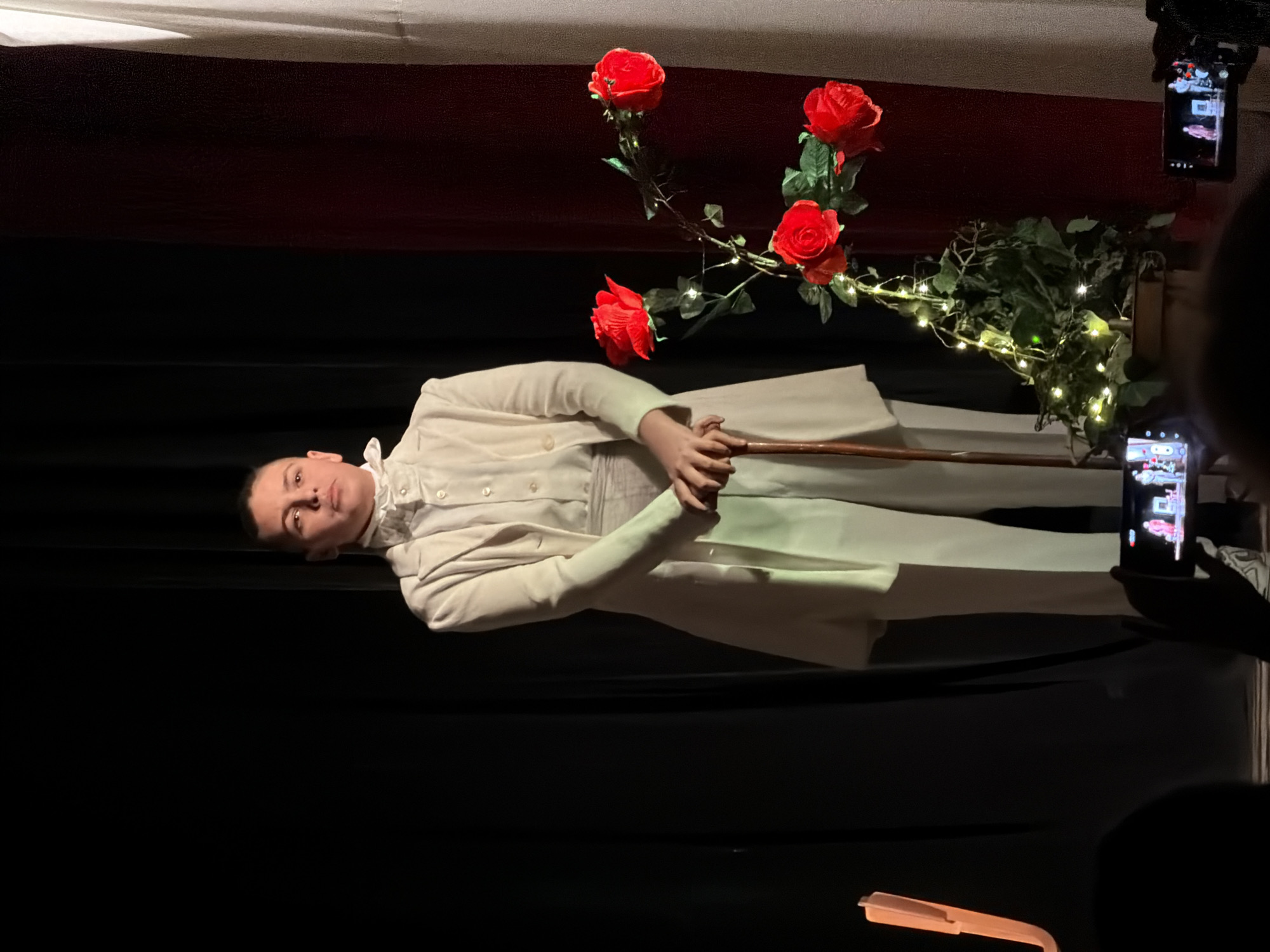 A person in a white suit holds a cane on a stage in front of a backdrop with red and white fabric, as well as a plant with red roses. Two people are taking photos of the scene. The photograph was taken in Pleven, Bulgaria.