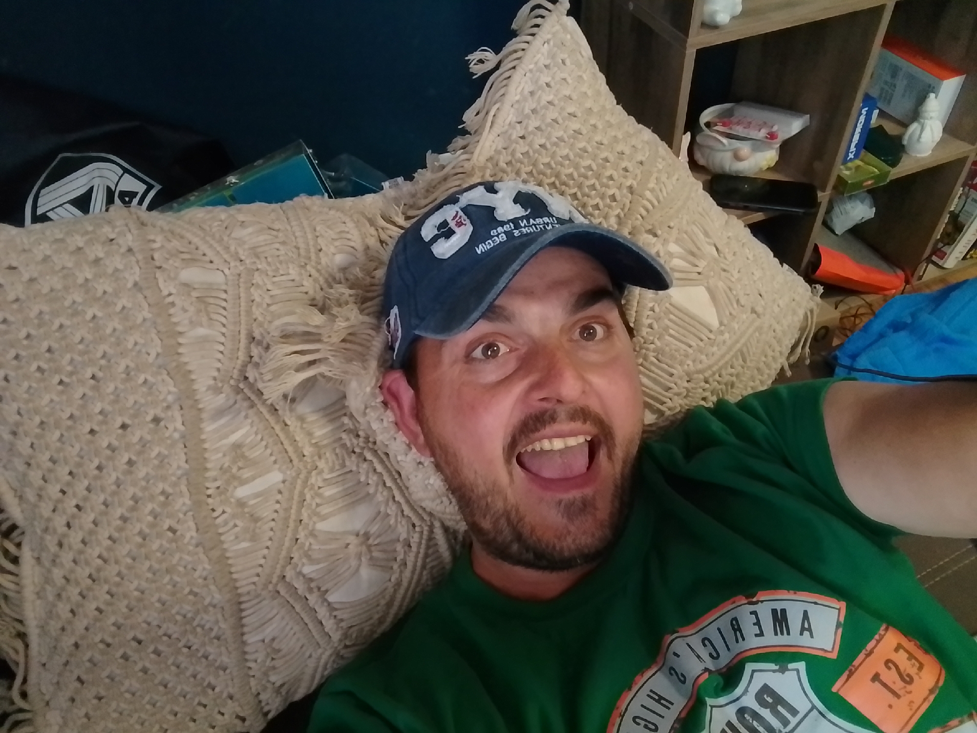 A selfie shows a white male lying down, wearing a blue baseball cap and a green t-shirt. He is smiling with his mouth open and looking at the camera. The background includes a macrame pillow, a shelf with various items, and a blue blanket. The location is not specified in the image, therefore the city of Wedel, Germany is not mentioned.