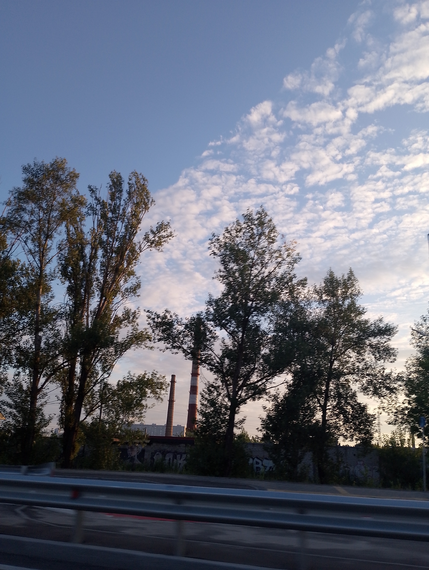 This is a photograph taken from a moving vehicle, likely a highway, showing a view of industrial chimneys against a backdrop of a partially cloudy sky. The time of day appears to be late afternoon or early evening, given the warm light and elongated shadows. The sky is a soft blue, with scattered white clouds that are illuminated by the sun.
In the middle ground, two tall, brick chimneys are visible. One is predominantly red and white striped, while the other is a reddish-brown brick. These chimneys suggest an industrial setting. Between the chimneys and the foreground, there is a low, weathered brick wall with graffiti. Trees with lush green foliage are scattered throughout the scene, some in the foreground and others behind the wall. The trees in the foreground are tall and have a somewhat sparse appearance.
The foreground is dominated by a highway guardrail, indicating the perspective is from a car or similar vehicle traveling along a road. The road itself is visible below the guardrail, with a wet surface reflecting the light, suggesting recent rain or the presence of dew. The overall mood of the image is somewhat serene, with the industrial elements softened by the natural beauty of the sky and trees. There is no visible text in the image, other than the graffiti on the wall, which is illegible. The location is identified as Kyiv, Ukraine.
