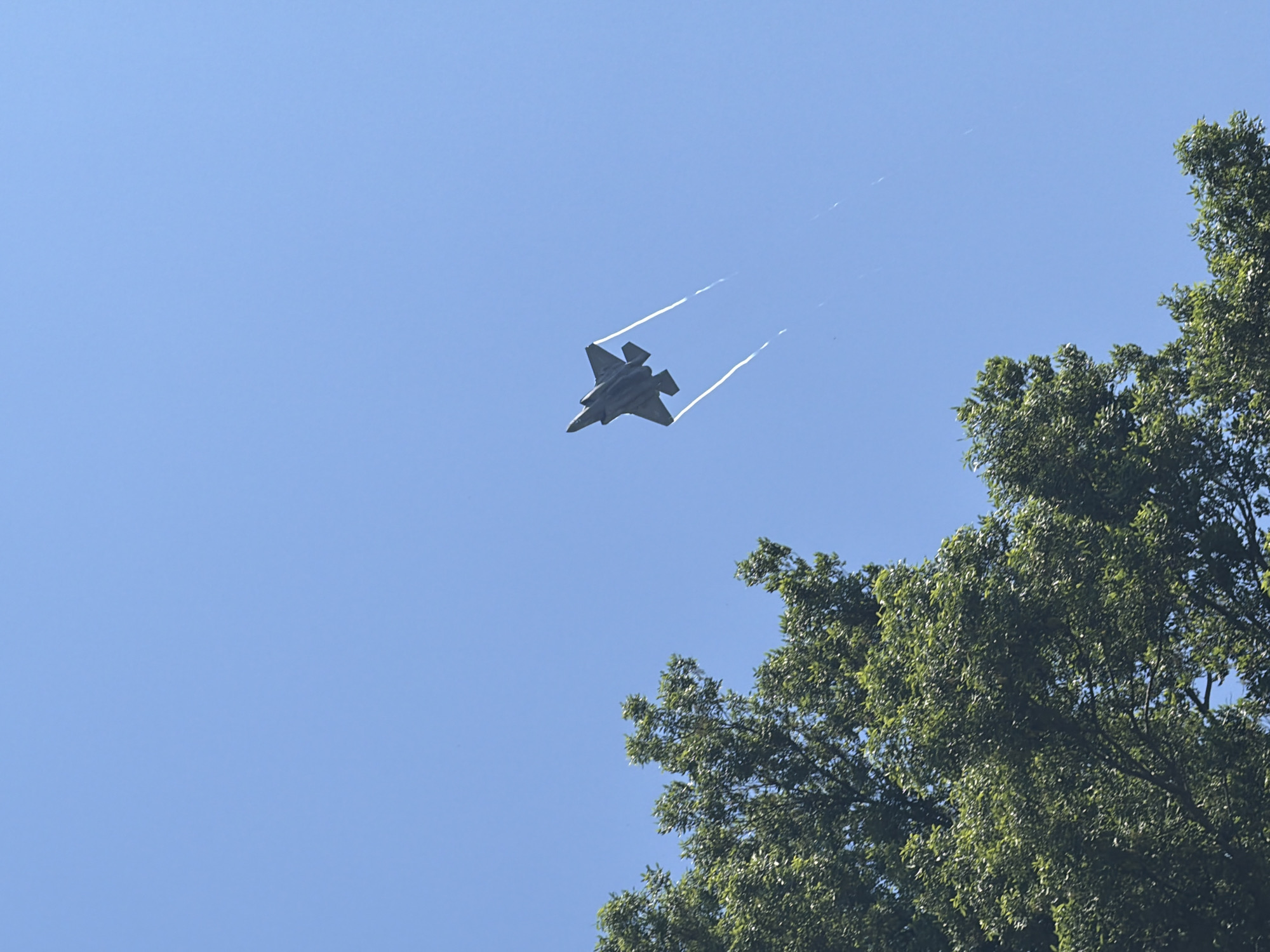 The image shows an F-35 fighter jet flying over the Naval Academy in Annapolis, Maryland. The jet is in the center of the frame, and contrails are visible behind it. In the lower right corner of the image, a tree is visible. The sky is a clear, light blue.