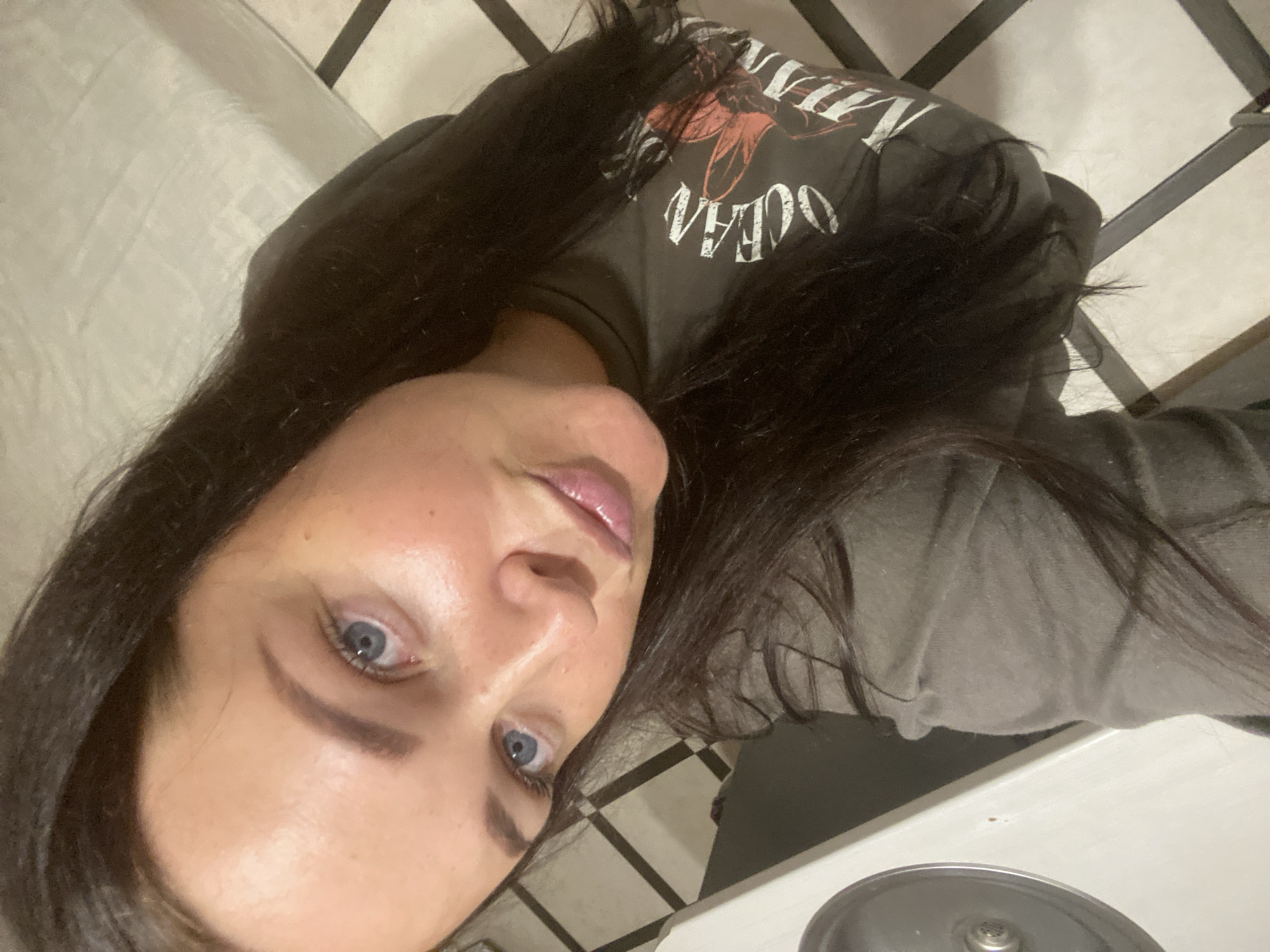 A woman with long dark hair and blue eyes is shown in a selfie. She is wearing a dark t-shirt with a graphic design. The background appears to be a tiled floor and a white surface, possibly a bed. The lighting is dim, suggesting an indoor setting. There is no indication of the location being in Sochi, Russia.