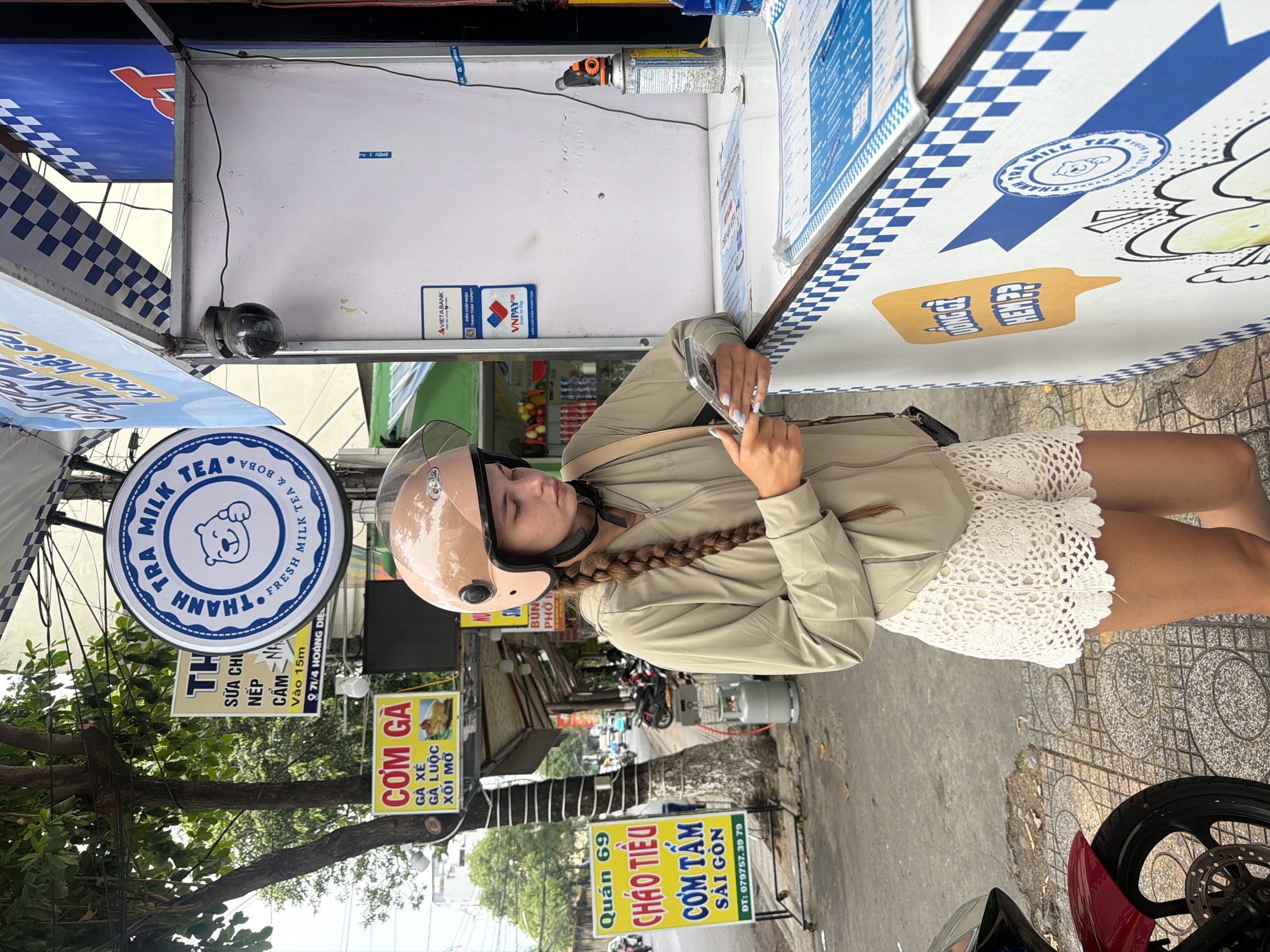 A young woman wearing a light beige jacket, white crochet shorts, and a pink helmet is standing on a sidewalk in Nha Trang, Vietnam, looking at her phone. She has a long braid that extends down her back. Behind her is a street food stall with several signs advertising food and drinks. One sign, in Vietnamese, reads "Com Ga" which translates to "Chicken Rice". Another sign, in English and Vietnamese, advertises "Thanh Tra Milk Tea" with a bear logo. The stall has a blue and white checkered pattern on its counter. A red motorcycle is parked to the left of the woman. The environment appears to be a busy street with other stalls and vehicles visible in the background.