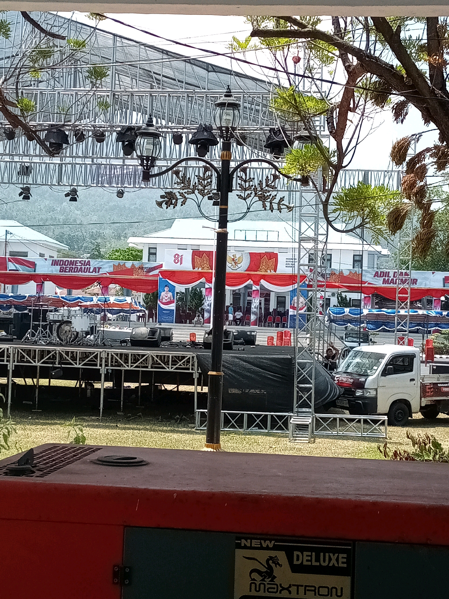 The image depicts an outdoor event setup in Bitung, Indonesia, likely a public gathering or concert. In the foreground, a large red generator unit is partially visible, with "NEW DELUXE MAXTRON" branding. Behind it, a raised stage structure with scaffolding and black tarping is set up. On the stage, musical instruments, including a drum set and speakers, are visible. A white truck with "smartive" branding and another truck with "SMART VIDEO" branding are parked near the stage. Red and white banners with Indonesian text and symbols, including "INDONESIA BERDAULAT" and "ADIL DAN MAKMUR," are displayed across the front of buildings in the background. The buildings appear to be official structures, possibly government or public offices, with a coat of arms and the number "81" prominently featured on one facade. Red chairs are arranged in front of the buildings. Trees with foliage partially obscure the view of the stage and buildings. The overall impression is of a prepared venue for a public event.