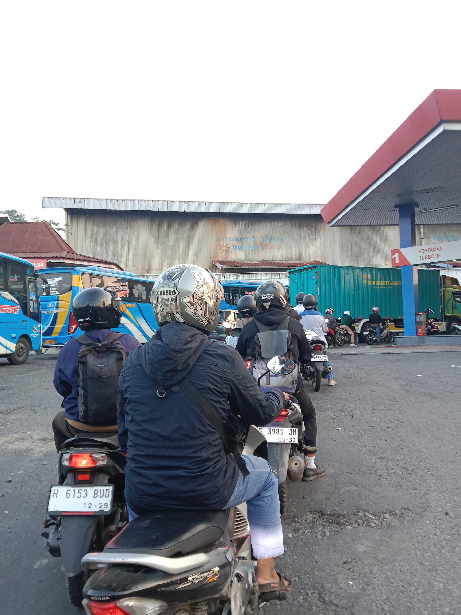 The image captures a scene at a Pertamina gas station in Semarang, Indonesia, with a focus on the motorcycles and buses present. In the foreground, several people are riding motorcycles, facing away from the camera. The rider in the immediate foreground wears a black jacket and a helmet with a decorative design. Behind them, another motorcycle carries two passengers, one wearing a blue jacket and the other a black jacket. Both have helmets on. The license plate of the first motorcycle reads "H 6153 BUD".
In the middle ground, a line of blue buses is visible, some with passengers inside. One bus has the text "TRANS SEMARANG" and the number "1.14" displayed. Further back, other motorcycles and people are seen at the gas station. A large teal shipping container is also parked nearby, with the text "PT. ALIA NASSETA LONGSTO" visible.
The gas station canopy, a prominent red structure, is visible on the right side of the image, with a "PERTAMAX TURBO 98" sign. The ground is paved with asphalt, showing signs of wear and tear. The sky is overcast and bright. The overall impression is of a busy urban environment, likely during daylight hours, with people engaged in daily activities such as transportation and refueling.