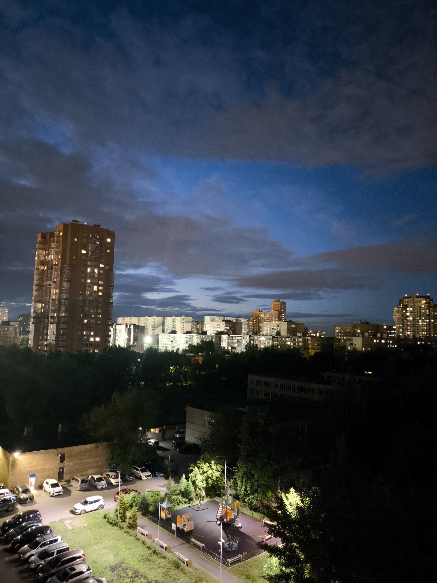 The image shows an urban landscape at night. In the foreground, there is a playground and a parking lot filled with cars. Beyond that, dense trees fill the middle ground, with several lit buildings visible in the background, including a prominent tall building with many illuminated windows. The sky is cloudy with patches of blue visible. The location appears to be a residential area.