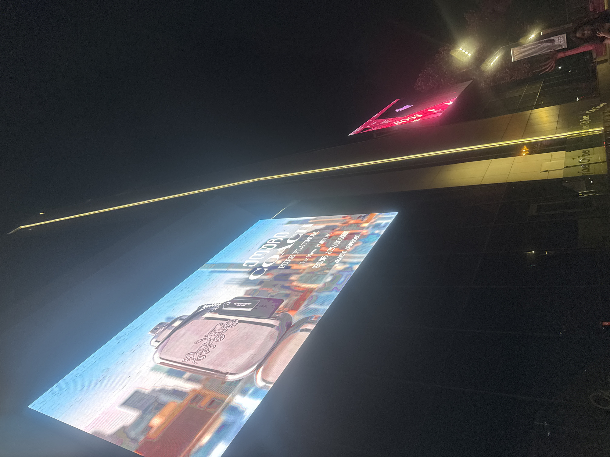 In the city of Tbilisi, Georgia, an advertisement for Coach perfume is displayed on a large LED screen mounted on a building. The screen shows a close-up of a perfume bottle with the Coach logo, set against a backdrop of a city skyline. Below the perfume bottle, text in Georgian reads "COACH PURE PLATINUM THE NEW PARFUM". Another advertisement, featuring the "BOSS" logo in pink neon, is visible on an adjacent building. The scene is illuminated by the bright lights of the advertisements and streetlights, creating a vibrant nighttime urban atmosphere.