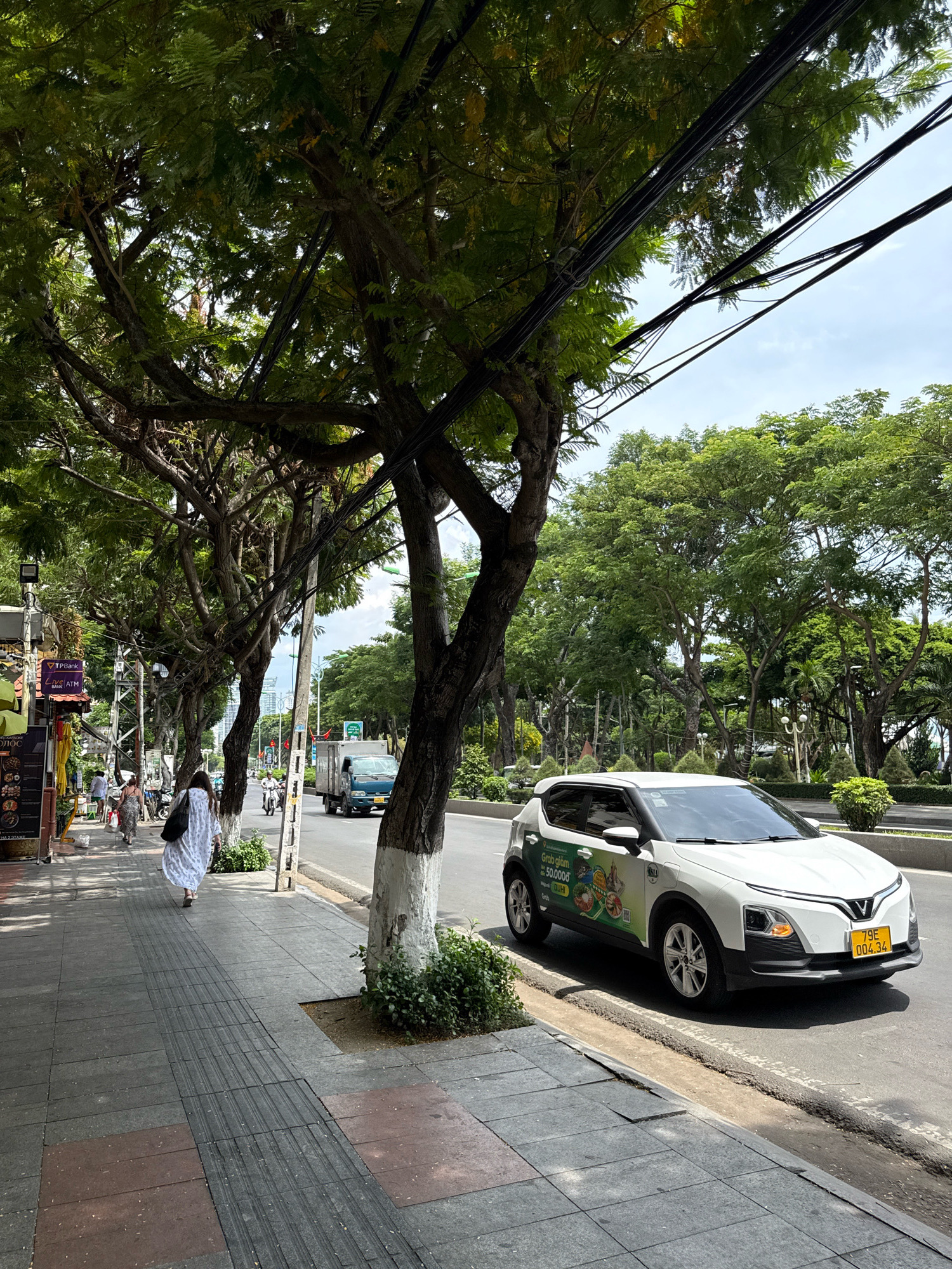 The image depicts a street scene in Nha Trang, Vietnam, likely during the day given the bright lighting and shadows. A sidewalk runs along the left side of the frame, populated by several individuals. A woman in a long, white, patterned dress walks away from the camera on the sidewalk, carrying a black bag over her shoulder. Other pedestrians are visible further down the sidewalk, some browsing shop displays or walking.
The street itself is paved and appears to be a multi-lane road. A white car with green decals on its side is parked or moving slowly on the right side of the road. The car has a Vietnamese license plate that reads "79E 004.34". A light blue truck is traveling in the same direction as the white car, further down the street.
The scene is characterized by lush greenery. Large trees with dense foliage line the sidewalk and the opposite side of the street, their branches stretching over the road and sidewalk. Power lines are strung between poles and trees, crisscrossing the upper part of the image. The sky is visible through the leaves, appearing bright and possibly partly cloudy.
The sidewalk features a tactile paving strip for visually impaired individuals, indicating a public thoroughfare. Buildings and shop fronts are visible along the left side of the street, with signage suggesting commercial activity. The overall impression is one of a busy urban environment with a significant presence of nature.