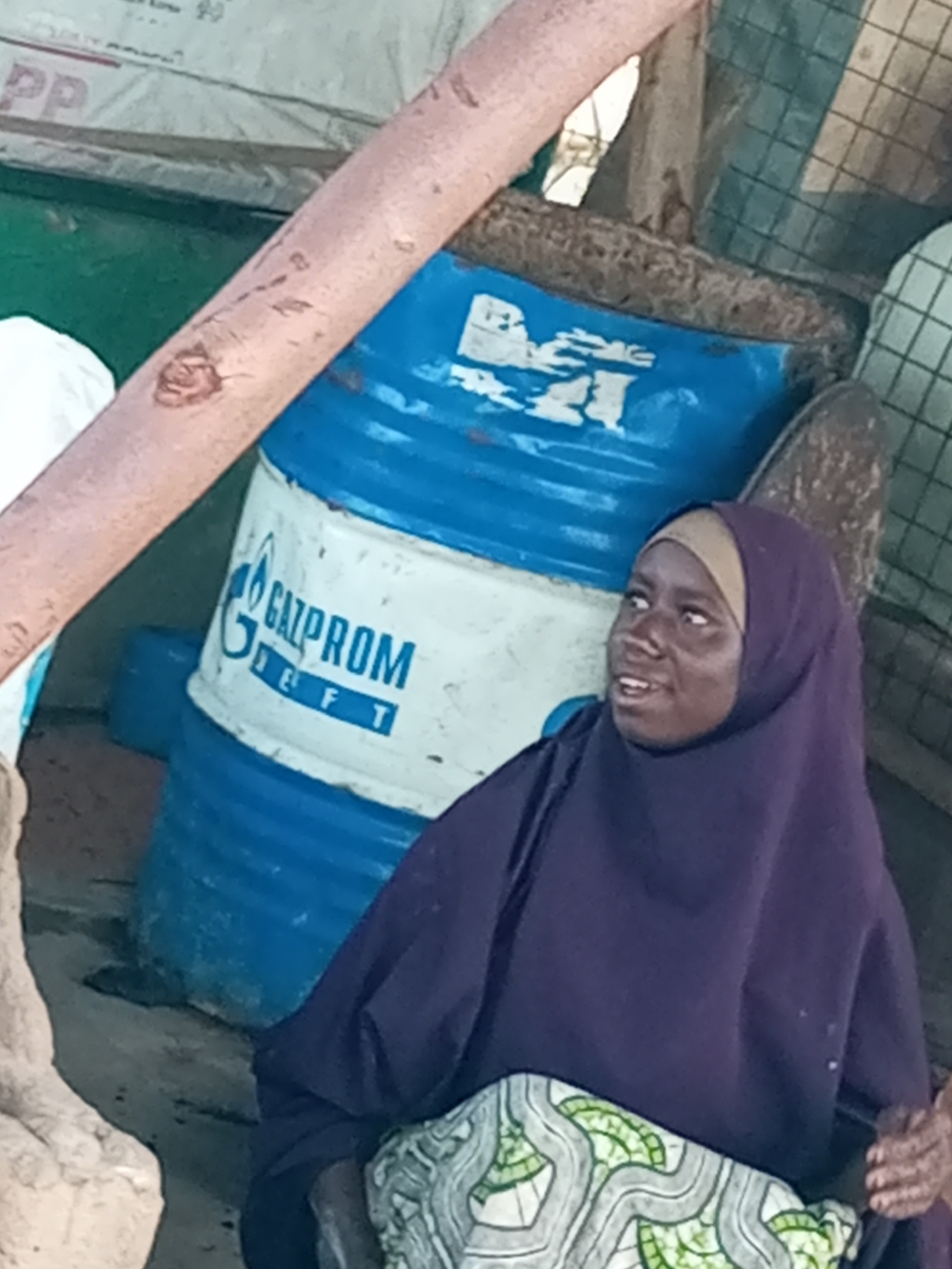 In Gombe, Nigeria, a person is seen wearing a purple hijab and a patterned skirt, sitting next to a large blue and white barrel. The barrel has the "GAZPROM" logo and "TEFT" written on it. The scene appears to be outdoors, possibly in a dusty or rural area, with a wooden structure and a rough, textured surface visible in the background. The lighting suggests it could be daytime. The person's expression is one of slight surprise or curiosity, with their mouth slightly open. The overall setting hints at a casual or informal gathering, or perhaps a moment of rest in a functional environment.