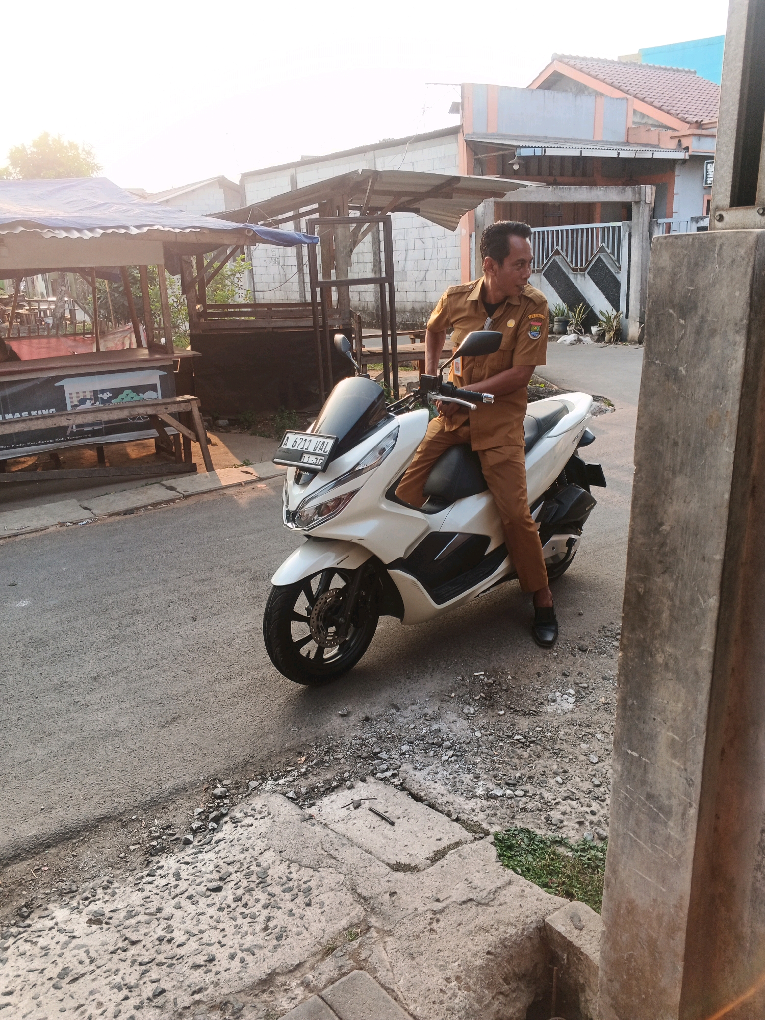 A man in a tan uniform sits on a white scooter on a paved street in Kadu, Indonesia. The man is looking to his left and has a pair of sunglasses tucked into his shirt pocket. Behind him are several market stalls and houses. The sky is bright and clear, suggesting it is daytime. The street is lined with concrete and some gravel.