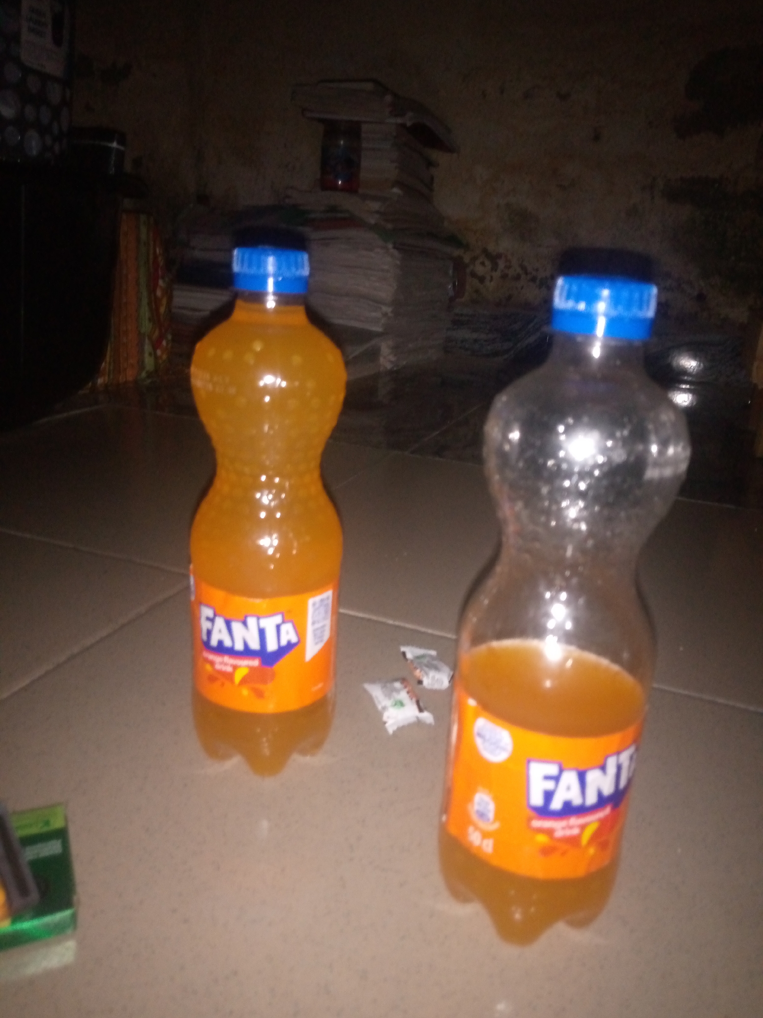 The image displays two plastic bottles of Fanta soft drink positioned on a tiled floor. One bottle, on the left, is full and unopened, while the bottle on the right is partially consumed. Both bottles have blue caps and feature the Fanta branding, with the orange liquid visible inside. Scattered on the floor near the bottles are a few small, white packets, possibly wrappers or sachets. The background is dimly lit and appears to be an indoor environment, with stacks of paper or books visible in the upper left quadrant and a dark, indistinct object to the far left. The lighting in the scene is low, creating shadows and highlighting the condensation on the bottles. The overall setting suggests a casual or domestic environment.