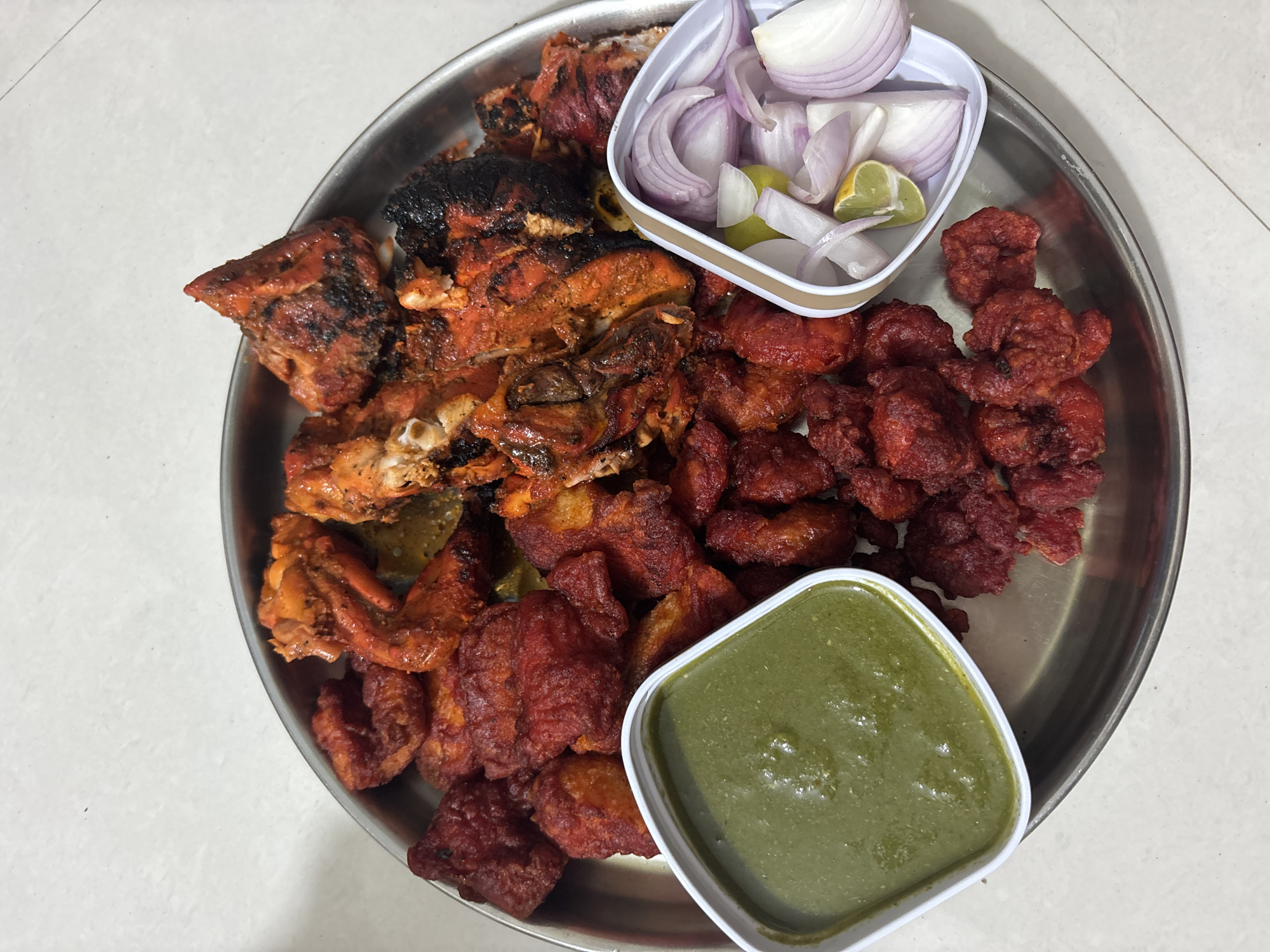 A top-down shot shows a round stainless steel plate holding two different types of cooked chicken dishes. On the left side of the plate are pieces of tandoori chicken, with some pieces appearing charred. On the right side, there are smaller, deep-fried chicken pieces coated in a reddish-brown sauce. A small white bowl in the upper right section contains sliced red onions and lime wedges. In the lower right section, there's another small white bowl filled with green chutney. The plate rests on a white tiled surface. The image appears to have been taken indoors, potentially in a restaurant or home setting, but the exact location in Ambernath, India, cannot be determined from the visual information provided.