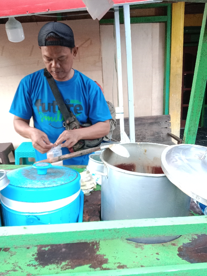 An Asian man in a blue "Future Smiles" t-shirt and black baseball cap stands behind a food cart. He is opening a small plastic bag. The cart displays a large metal pot with a ladle inside and a blue beverage cooler. The cart's surface is painted green. In the background, there is a building with green, yellow, and white trim. A lightbulb hangs above the man.