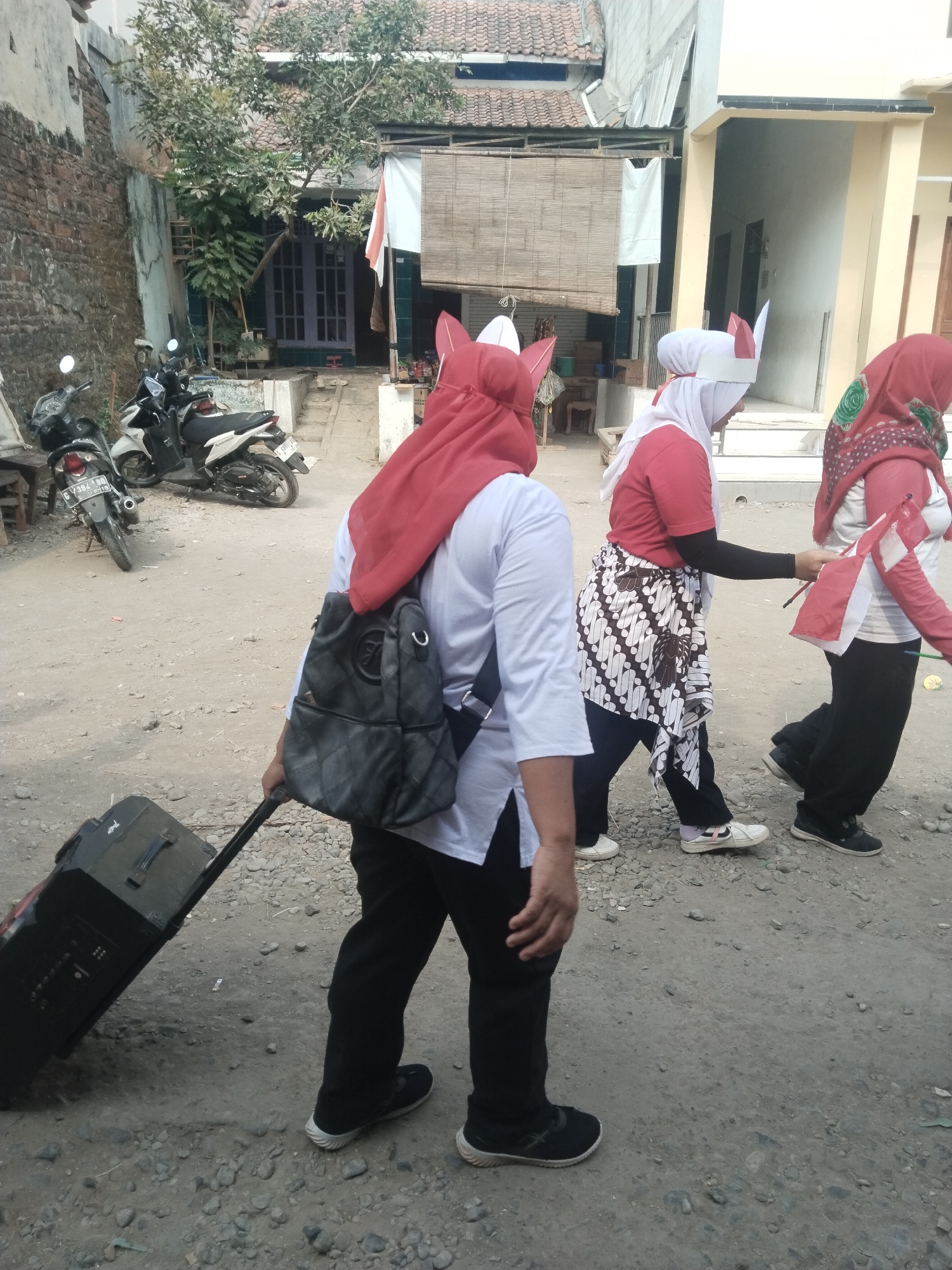 In Jembayat, Indonesia, three women are walking along a dusty street in front of buildings. The woman in the foreground, wearing a red headscarf and a white shirt, is pulling a large black rolling suitcase. She has a backpack on her shoulders. To her right, another woman in a red shirt and white headscarf wears a batik-patterned sarong over black pants and is holding a small Indonesian flag. A third woman, also wearing a red headscarf and a white shirt, is walking slightly ahead of her. Motorcycles are parked on the left side of the street. The environment appears to be a residential area with a simple, somewhat unkempt appearance.