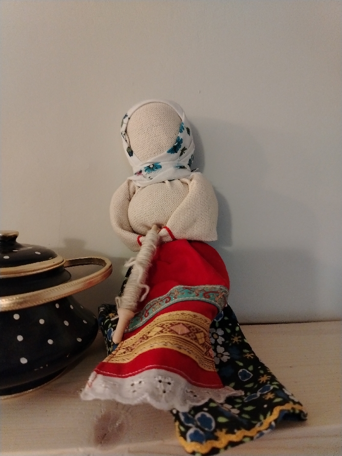 The scene displays a traditional Russian-style fabric doll and a lidded pot, resting on a white surface against a light-colored, plain wall. The doll, positioned centrally to the right, lacks facial features, composed of light-colored fabric for its head and torso. It wears a white headscarf with blue and green floral patterns, a light-colored long-sleeved top, and a multi-layered skirt. The skirt features an inner dark fabric with blue and yellow patterns, a red layer with a turquoise, gold, and red embroidered band, and an outer white layer with eyelet lace trim. The doll holds a wooden spindle wrapped with light-colored fiber in its arms. To the left, partially obscured, is a black lidded pot adorned with white polka dots and a gold-colored metallic handle. This depiction of traditional craftsmanship is located in Lezhnevo, Russia.