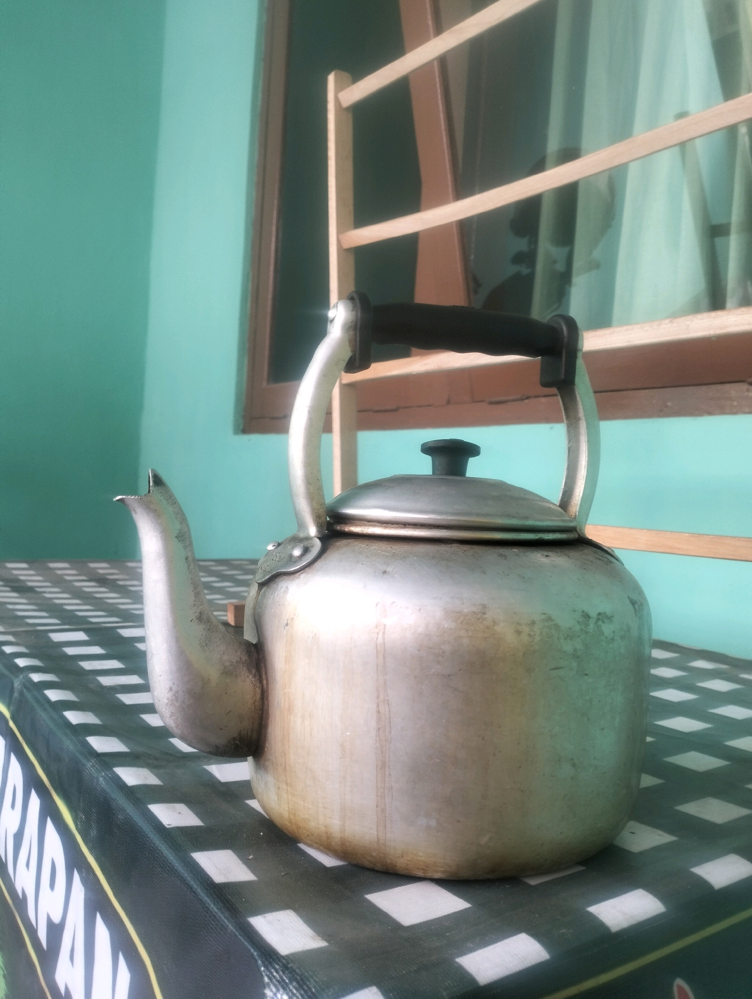 The image depicts a traditional aluminum kettle placed on a table covered with a black and white checkered tablecloth. The kettle is made of brushed aluminum and has a black handle. It has a spout and a lid with a knob. The background features a turquoise wall and a window with wooden frames and horizontal wooden bars. Behind the bars, sheer curtains are visible. A portion of what appears to be a banner or large piece of fabric with white lettering and a yellow stripe is visible at the bottom left of the image, partially obscured by the kettle. The scene is lit by natural light, creating soft shadows. The overall setting suggests an indoor environment, possibly a home or a simple eatery, in Kembaran, Indonesia, given the architectural style and the presence of what looks like local textiles. There are no people visible in the image.