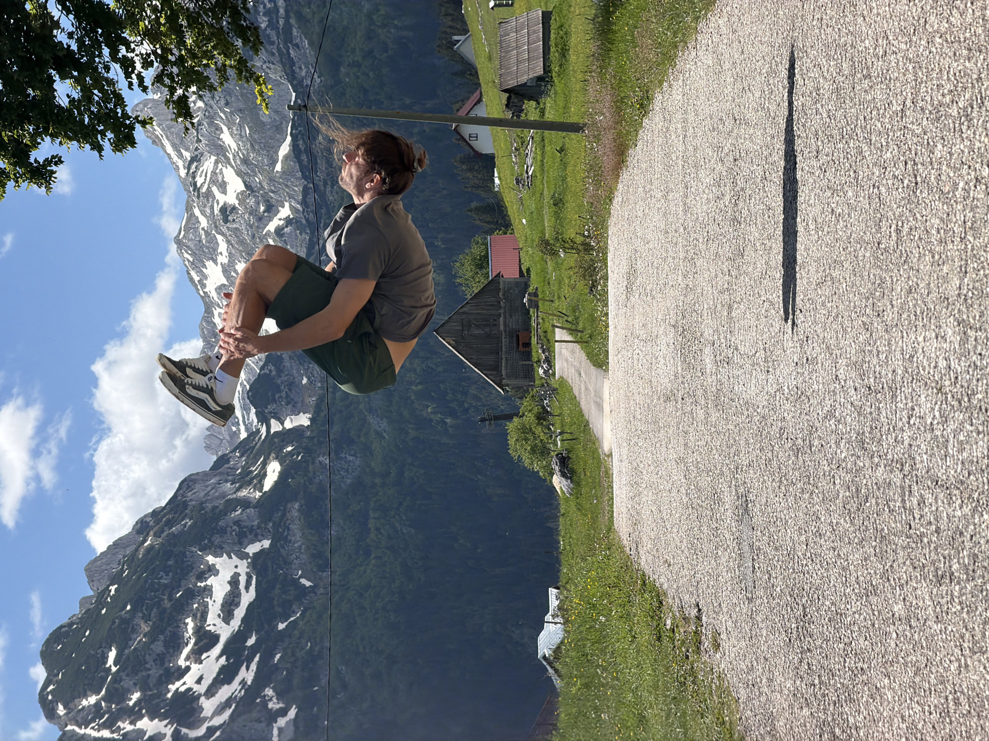 This image depicts a dynamic outdoor scene in Pitomine, Montenegro, characterized by a vibrant natural landscape. The central subject is a young man captured mid-air, performing a jump or leap. He is positioned with his legs bent upwards towards his chest and arms close to his body, indicating active movement. He wears a grey t-shirt, dark green shorts, and light-colored sneakers with black stripes. His light brown hair, partially tied back, appears windblown, suggesting motion or a breeze. His gaze is directed upwards and to the right, towards the sky.
The background features majestic, rugged mountains, parts of which are covered with patches of snow and dense green forests. A clear blue sky with scattered white clouds extends above the peaks. In the mid-ground, nestled within a green valley, are several traditional-style wooden houses or cabins with dark roofs, surrounded by fields.
In the foreground, a textured, light-colored gravel path extends along the right side of the frame. A long, thin shadow is cast across this path, indicating strong sunlight and suggesting the time of day is late morning or early afternoon. A tree branch with lush green leaves occupies the upper left corner, framing the scene. The overall impression is one of energetic human activity set against a beautiful, sunny, and mountainous backdrop. There is no visible text in the image.