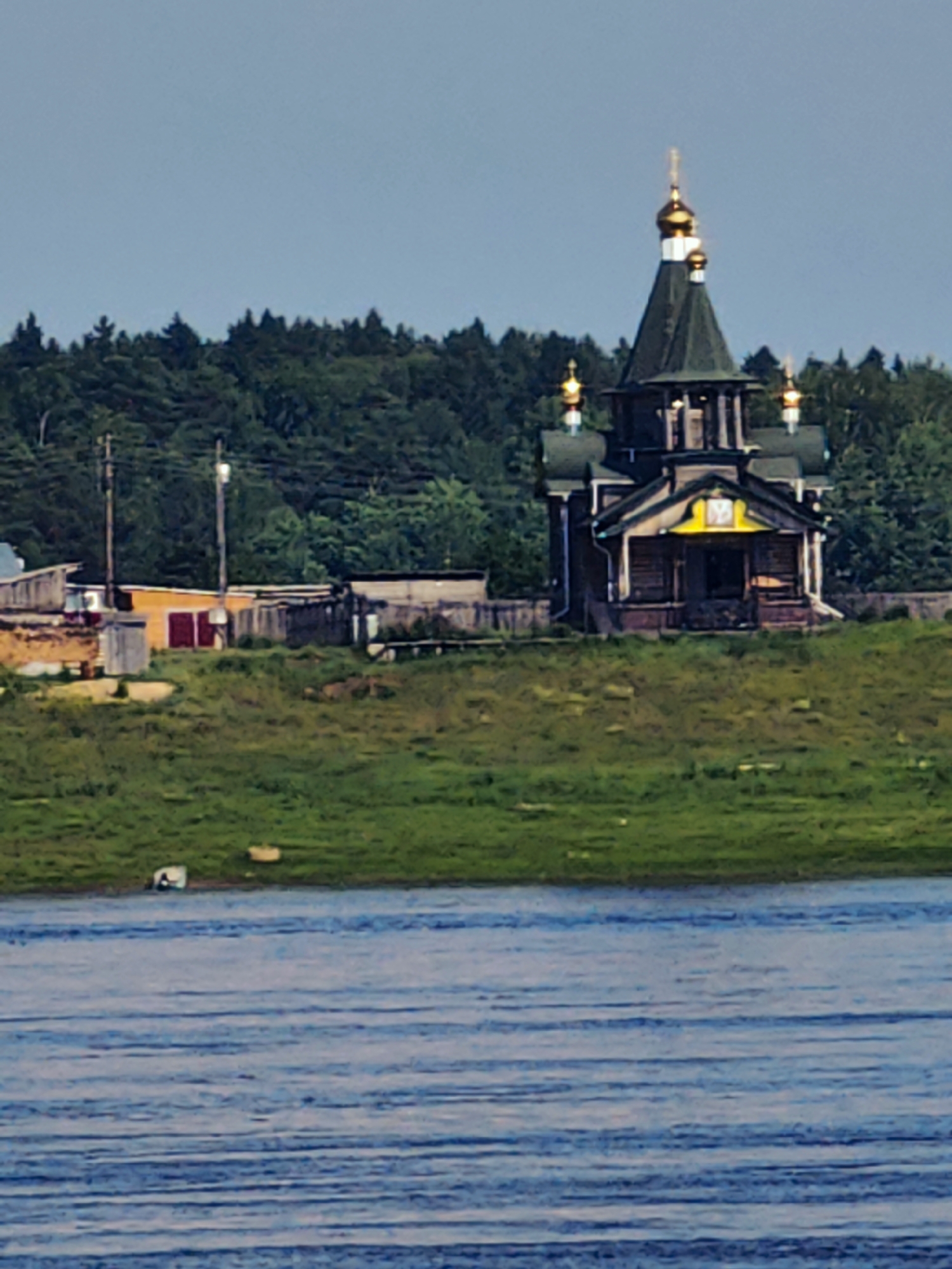 An Orthodox church with a green roof and golden domes stands on a grassy hill overlooking a body of water. The church is a wooden structure with a yellow emblem above the entrance. To the left of the church, there are several smaller wooden buildings and fences, suggesting a small village or settlement. The background is dominated by a dense forest of trees. The water in the foreground appears calm with visible ripples. The sky is a pale blue.