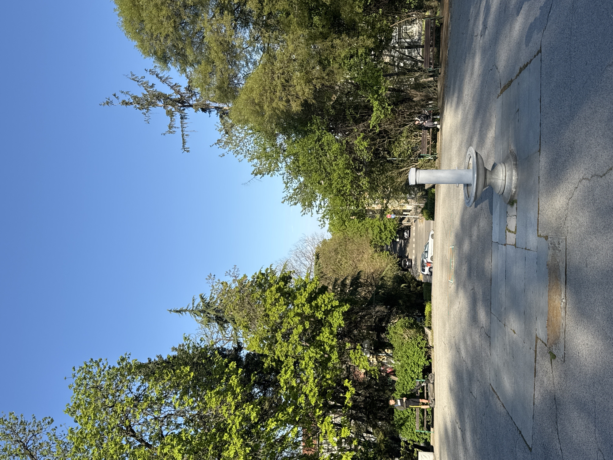 The image shows a park scene in Plovdiv, Bulgaria, during daylight. A gray drinking fountain is situated on a paved area. Trees line the perimeter of the scene, with a clear blue sky visible above. A street with parked vehicles can be seen beyond the trees. A bench with two individuals is visible next to the trees on the right.