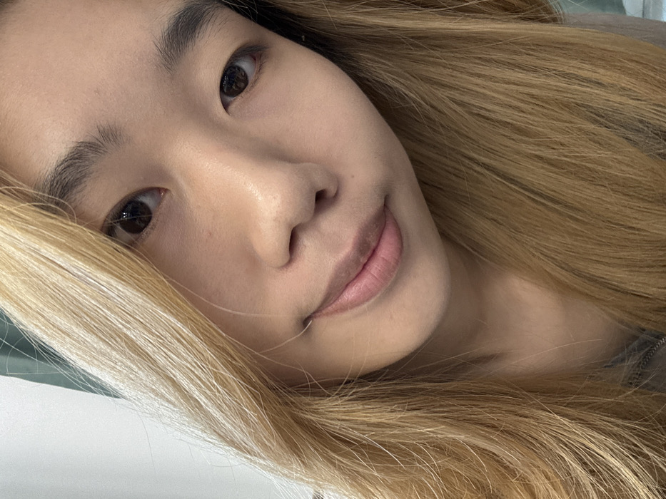 The image displays a close-up, slightly angled portrait of an East Asian female, depicted indoors within the city of Nan'an District, China. The subject faces the camera directly with a subtle, closed-mouth smile. Her long, wavy hair, colored light blonde, frames her face and extends downwards. Her facial features include dark eyes and dark, well-defined eyebrows. Her complexion appears even and smooth, possibly enhanced with minimal makeup. A portion of a gray garment, consistent with a hooded sweatshirt or sweater, is visible around her neck and upper chest. The background is out of focus, consisting primarily of a light-colored surface, possibly a wall, with a faint darker, greenish-gray element visible on the upper left. Illumination is soft and even, suggesting an artificial indoor light source. No other individuals or prominent objects are discernible in the scene.