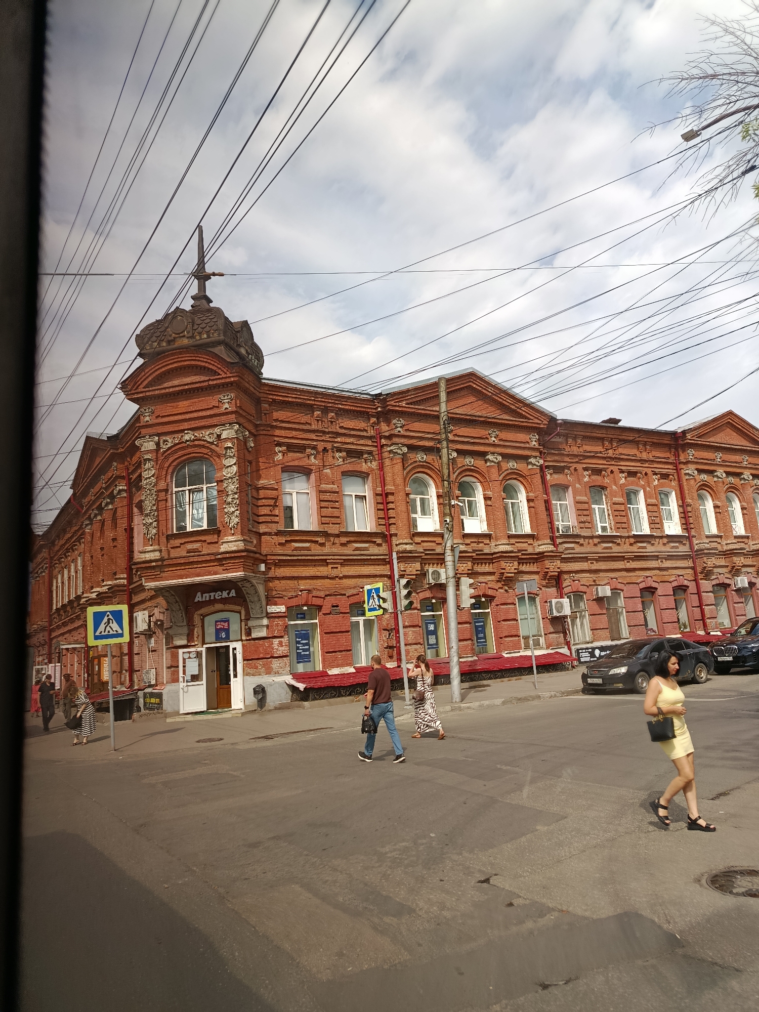 The image depicts a street scene in Saratov, Russia, featuring a prominent red brick building with ornate architectural details. The building is a multi-story structure with numerous arched windows and decorative elements. A sign above an entrance reads "АПТЕКА," indicating a pharmacy.
Several people are visible walking on the sidewalk and crossing the street. On the right side of the frame, a woman in a yellow dress is crossing the street, carrying a black purse. Further down the street, a man in a maroon t-shirt and jeans is walking away from the camera, holding a camera. Several other individuals are seen walking on the sidewalk in front of the building.
Cars are parked along the street and driving by. Two dark-colored sedans are visible on the right side of the image. Overhead, a complex network of electrical wires crisscrosses the sky, obscuring parts of the building and the sky. The sky is partly cloudy. Yellow pedestrian crossing signs are present at intersections. The overall scene suggests a typical day in a city environment.