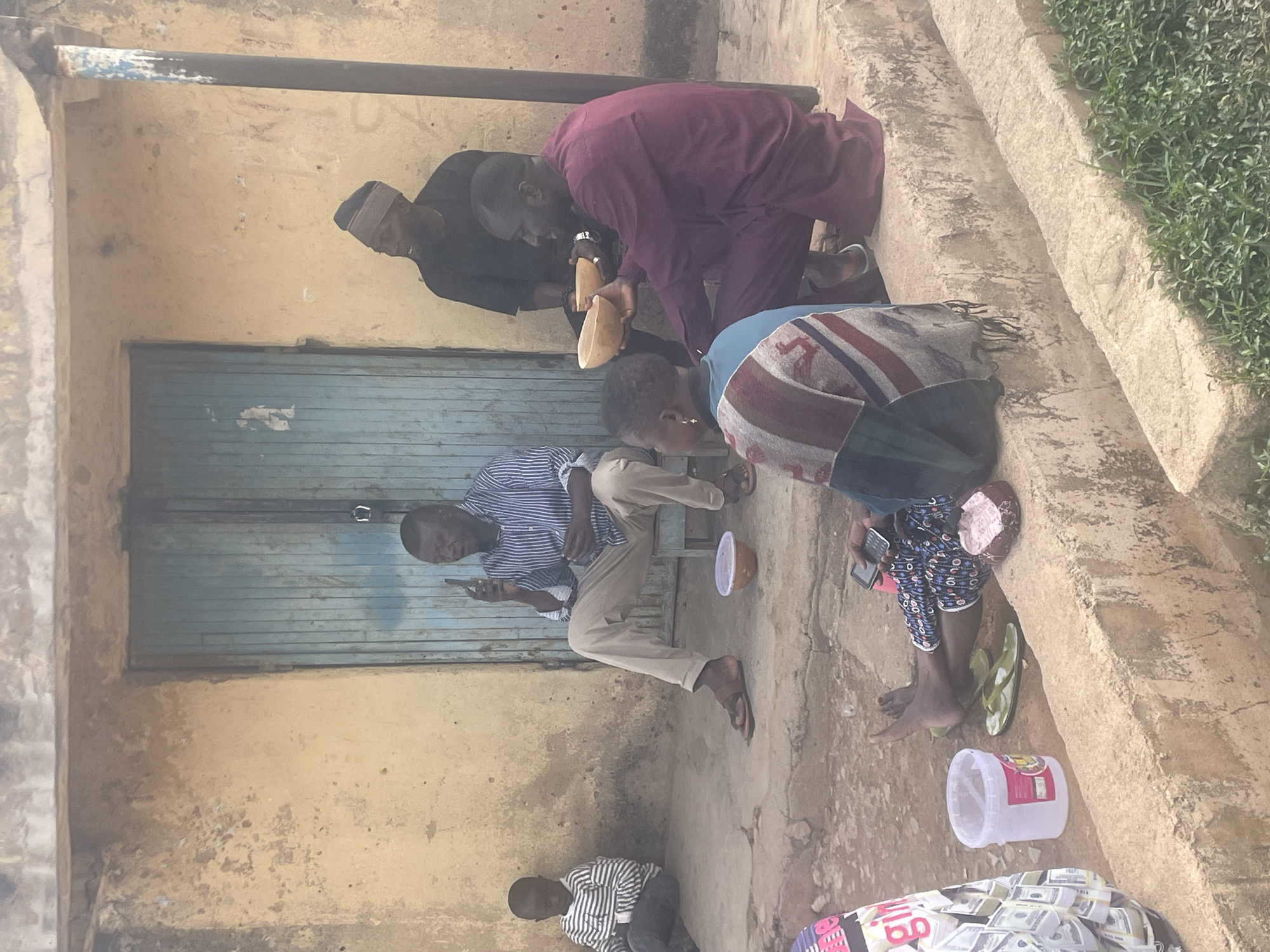 The image depicts a group of people in an outdoor setting. Three men are seated or kneeling on a set of stone steps, while another man is seated on a metal stool in front of a building with a closed, blue metal shutter. One man is looking at a phone in his hand. Another man is holding what appears to be a wooden object. A child is sitting on the ground. Near the edge of the steps, there is a clear plastic container and a patterned bag that appears to contain paper currency. The setting appears to be a street scene with worn, aged structures and greenery visible in the background. The location is Jos, Nigeria.