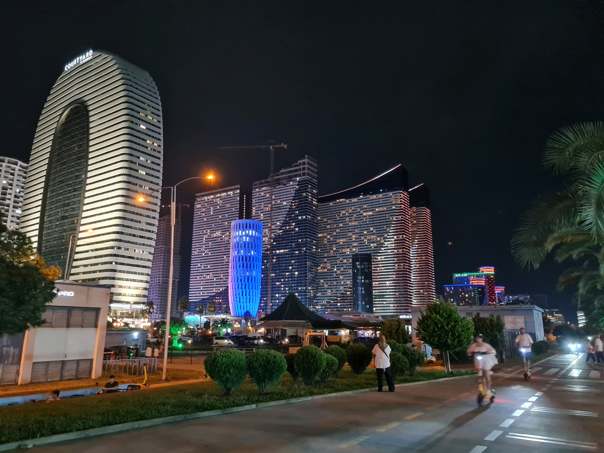 This image depicts a nighttime scene in Batumi, Georgia, likely along the Boulevard. Dominating the left side of the frame is the striking Courtyard by Marriott Batumi hotel, a tall, curved skyscraper with white horizontal banding and a large arched opening. To its right are several other modern skyscrapers, illuminated with numerous windows and some featuring colorful exterior lighting, particularly a prominent blue-lit cylindrical building. In the foreground, a paved pathway or road is visible, with several people riding electric scooters. One woman in a white top and dark pants stands on the side of the pathway, facing away from the camera. There are green bushes and trees lining the pathway, and further back, tents or canopies suggest an outdoor dining or gathering area. A street lamp with an orange light is visible in the middle left of the frame. The overall impression is of a lively urban environment at night, characterized by contemporary architecture and recreational activities.