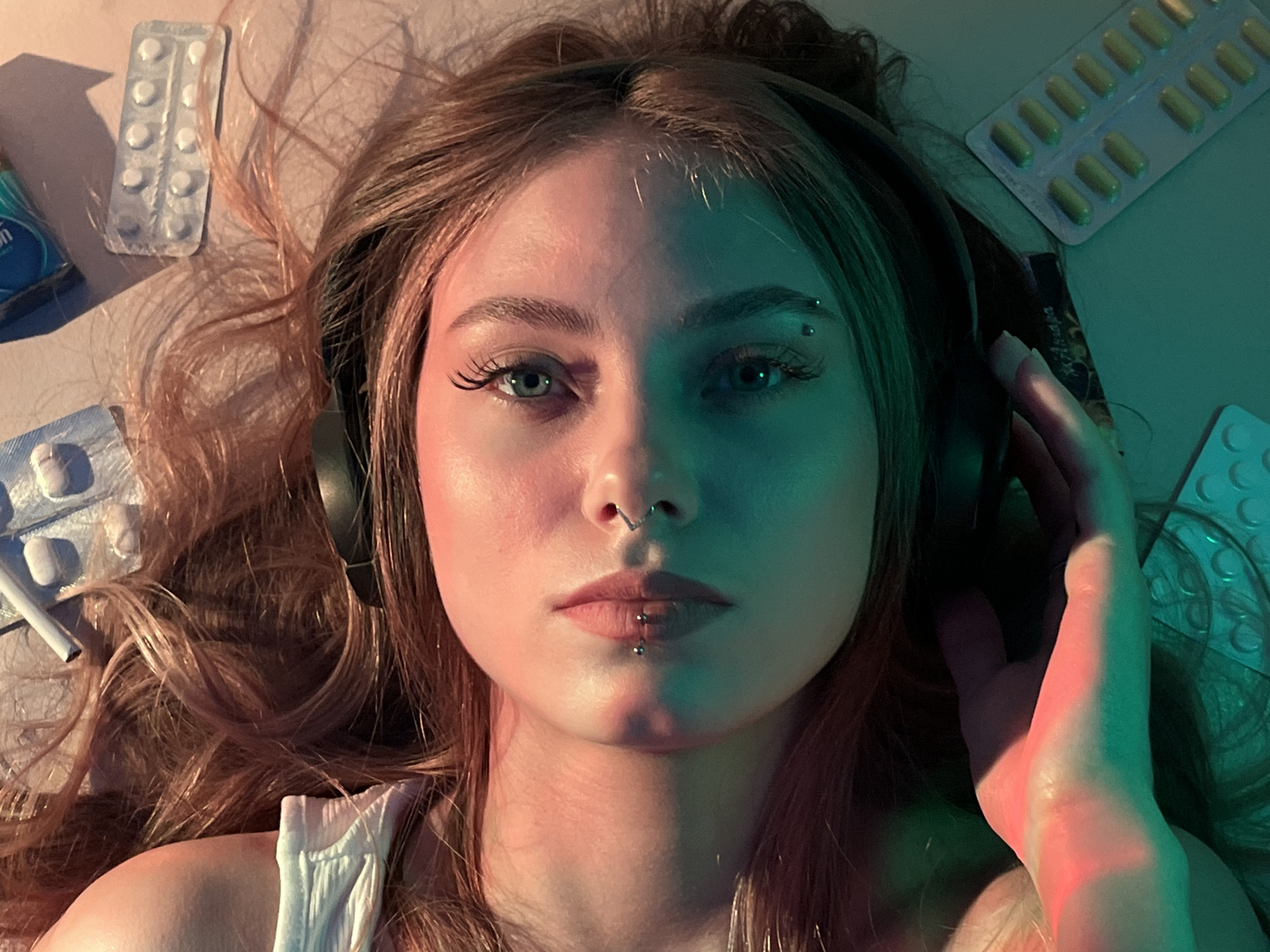 A young woman lies on her back with her head centered in the frame, wearing headphones. To her left, there are two blister packs of white pills and a cigarette. To her right, there is a blister pack of yellow pills. The woman has a nose ring, a lip ring, and an eyebrow piercing. She is wearing a white tank top. Her right hand is raised near her head. The lighting in the scene is predominantly green and pink. This scene is not necessarily in Фоки, Russia. The claim that it is located there is unfounded.