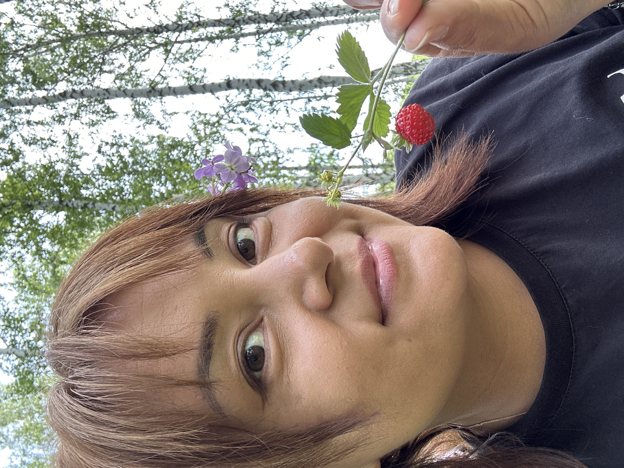 An outdoor medium close-up photograph shows a woman with medium-length brown hair and a black t-shirt holding a wild strawberry and flower near her face. The woman is looking slightly to her left, with her right eye visible and her left eye partially visible. She has a gentle smile on her face. A small purple flower is tucked behind her right ear, and a stem with green leaves and one red strawberry is held up to her mouth. The background consists of tall, slender birch trees with white bark and green foliage against a bright sky. The overall impression is one of a spontaneous moment captured in nature.