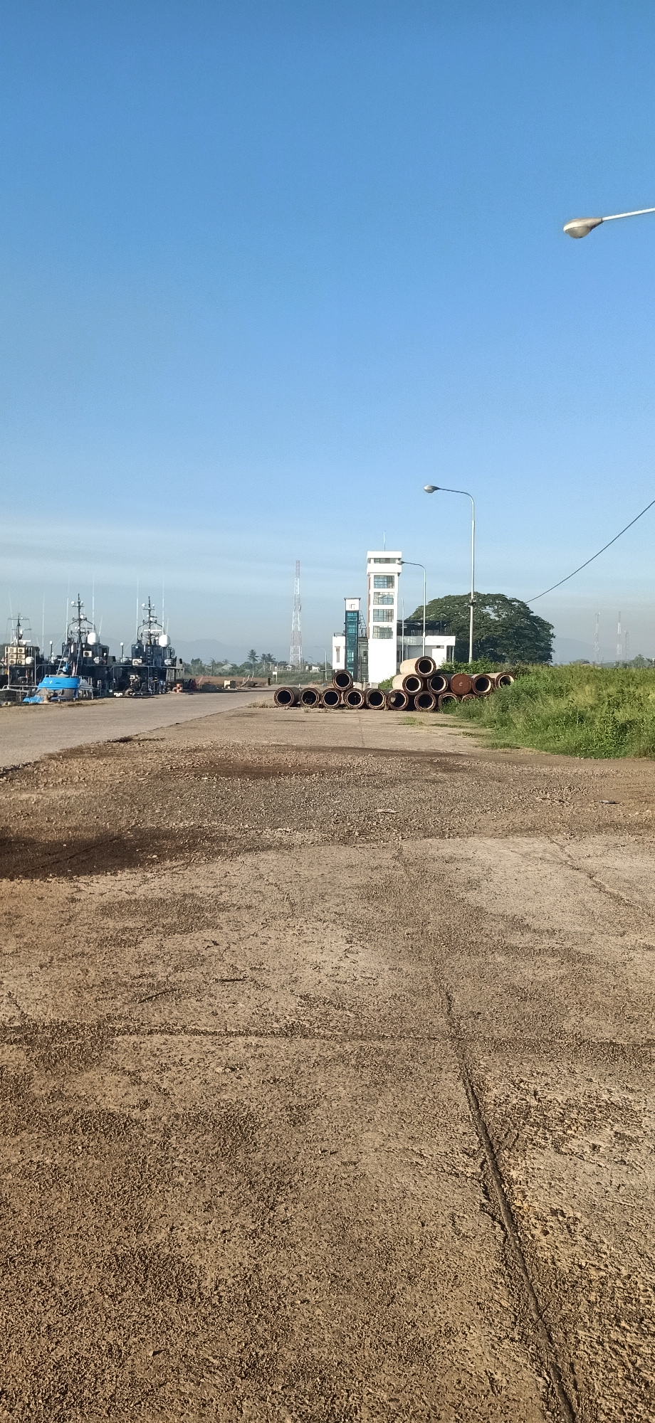 This is a vertical, eye-level shot of a harbor or industrial area in Keudekruenggeukueh, Indonesia. The foreground is dominated by a rough, cracked concrete surface. In the midground, to the left, several dark-colored ships with tall superstructures are docked. To the right of the ships, a collection of large, cylindrical pipes are stacked on the ground. Further back, a white building with multiple windows and a tower-like structure stands against a backdrop of green vegetation and distant hills. A tall, slender radio tower is visible to the left of the building. The sky above is a clear, bright blue. A street lamp is visible in the upper right quadrant of the image. The overall scene suggests a functional port or industrial zone.