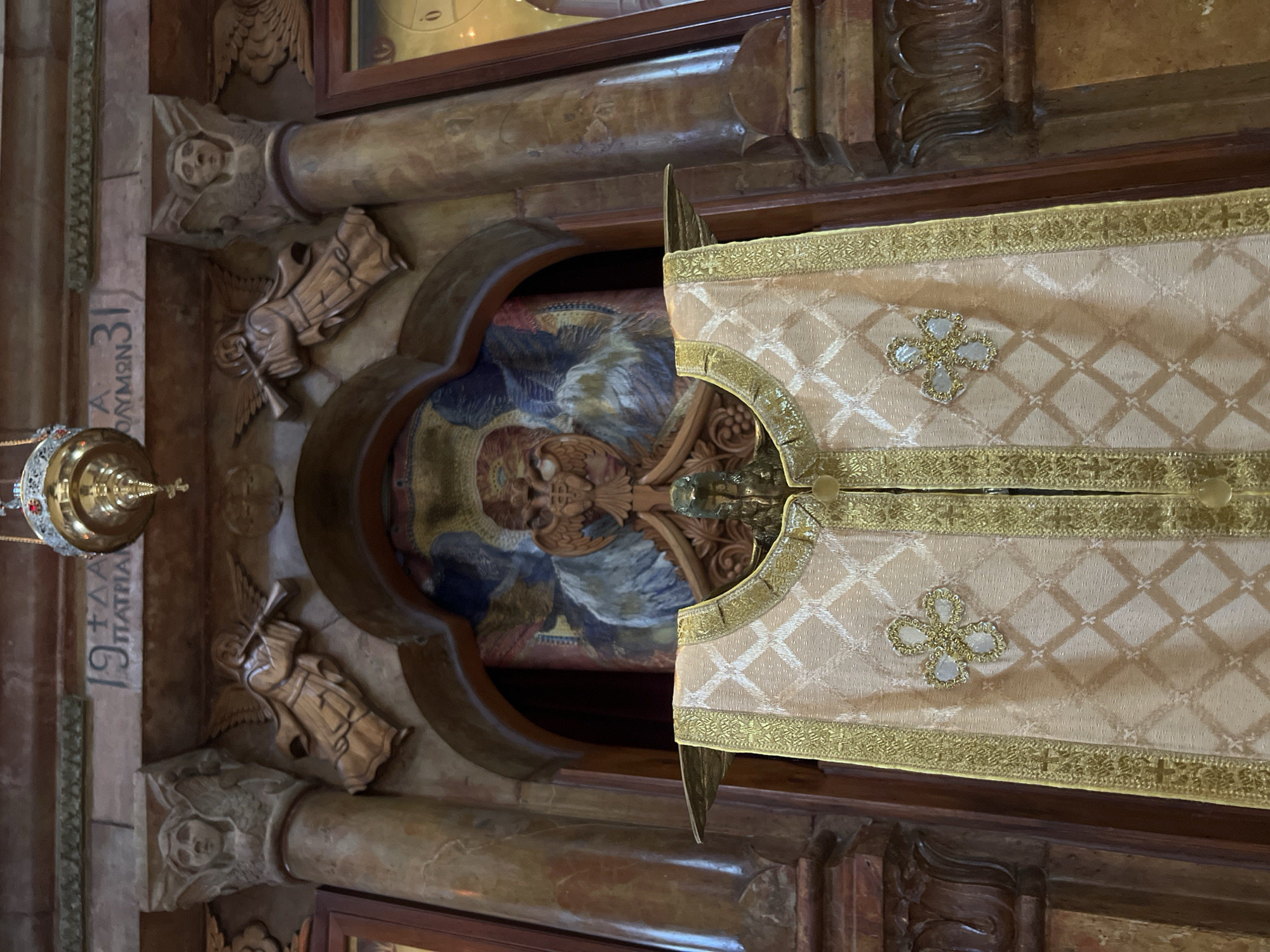 In an interior religious setting, a close-up shot focuses on a ceremonial vestment, likely a cope or chasuble, made of light-colored fabric with a diamond-shaped pattern and gold trim. It is draped over a stand with brass-like wing-shaped extensions. The vestment features ornate gold embroidery on the front, including four-petal floral designs. Behind the vestment, a mosaic depicts a winged figure with multiple faces, possibly an angel or seraph, rendered in shades of blue, red, and gold. The background includes carved stone pillars with angelic figures and inscriptions. To the left and right, marble columns flank the central mosaic. Above, a brass lamp hangs from the ceiling. The overall impression is one of religious ceremony and artistic craftsmanship.
