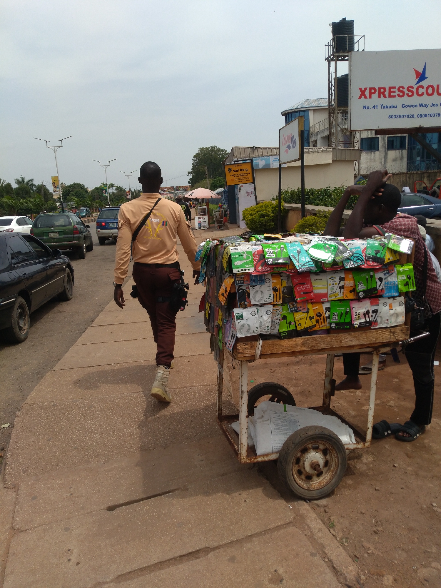 A man in an orange shirt and dark pants walks along a sidewalk in Jos, Nigeria. He is walking away from the camera and is seen from the back. To his right, a vendor is displaying mobile phone accessories on a cart. The cart is laden with various items, including earphones, chargers, and data cables, all packaged in colorful plastic. The vendor, partially visible, leans on the cart, resting his head on his hand. In the background, traffic can be seen on a road, with several cars and a green minivan. Buildings line the street, including a tall structure with water tanks on top and a prominent sign for "XPRESSCOUT." The sky is overcast. The scene depicts a typical street vendor setting in a Nigerian city, highlighting the sale of mobile accessories.