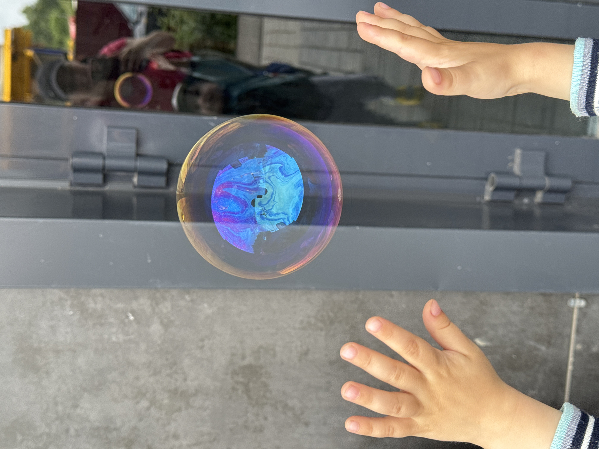 In Izhevsk, Russia, two children's hands are visible in the foreground, reaching towards a large, iridescent soap bubble. The bubble floats in front of a building with a grey, textured wall on the left and a reflective surface on the right. The reflective surface shows a mirror image of a person in a dark hat and pink shirt, holding a phone to take a photograph, and a child standing behind them. The scene appears to be outdoors, with hints of greenery in the background reflection. The overall impression is one of playfulness and a captured moment of childhood joy.