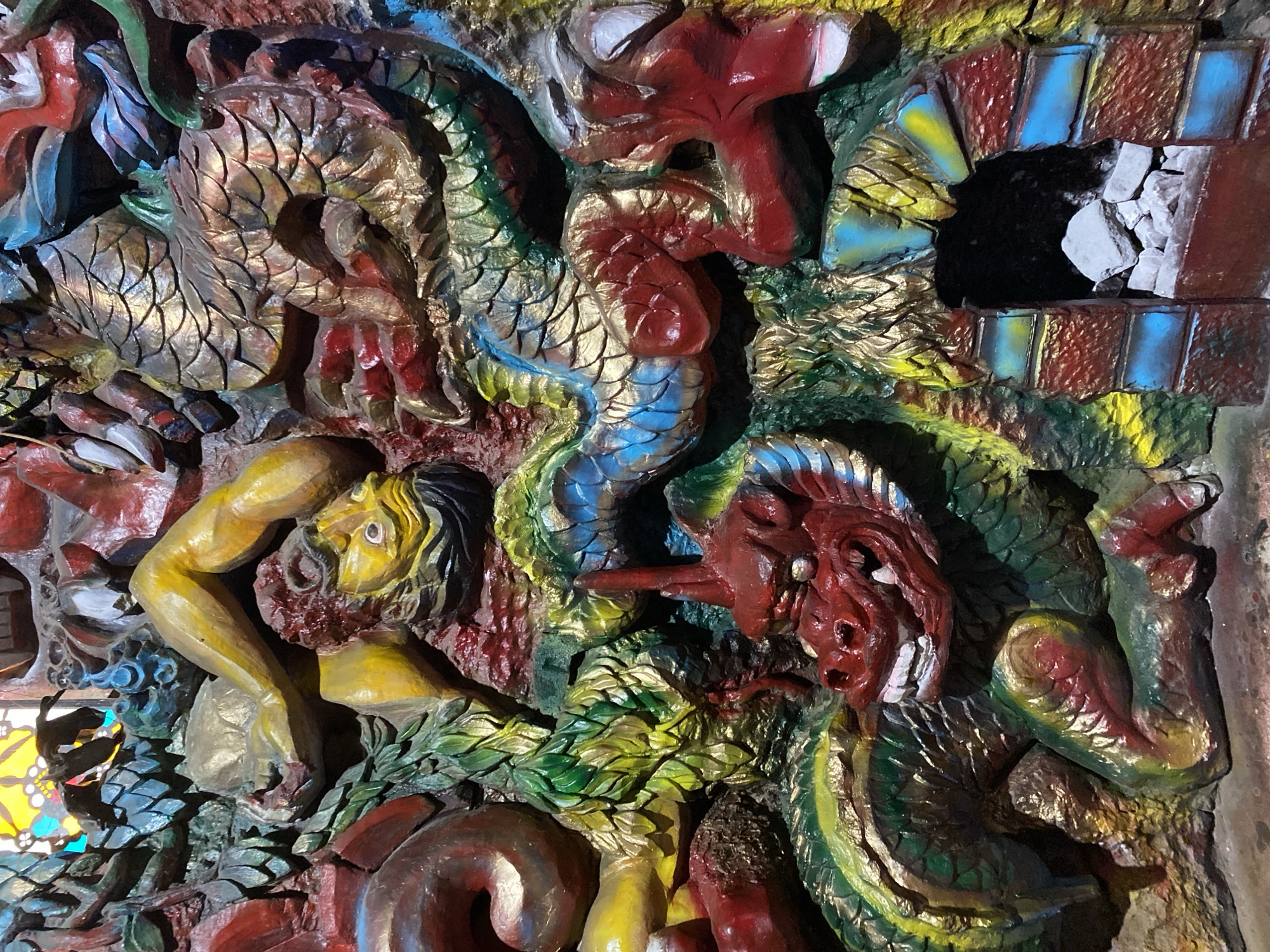 This is a close-up, eye-level, medium shot of a colorful, heavily detailed bas-relief sculpture. The sculpture depicts a mythological scene, possibly a dragon or serpent-like creature, intertwined with human-like limbs and faces. The creature's body is covered in scales painted in shades of gold, green, blue, and red. Several human-like limbs, rendered in yellow and red, are visible, suggesting a struggle or interaction. One human face, with yellow skin and red hair, is depicted with wide eyes and an open mouth. Another prominent feature is the head of a red dragon-like creature with sharp teeth and horns, positioned in the foreground and looking towards the viewer. To the right, there is an arched opening resembling a cave or doorway, with stacked stones inside. The sculpture is densely packed with elements, with no discernible background beyond the artwork itself. The lighting is somewhat uneven, highlighting the texture and colors of the sculpture. The location is not specified in the image or crops.