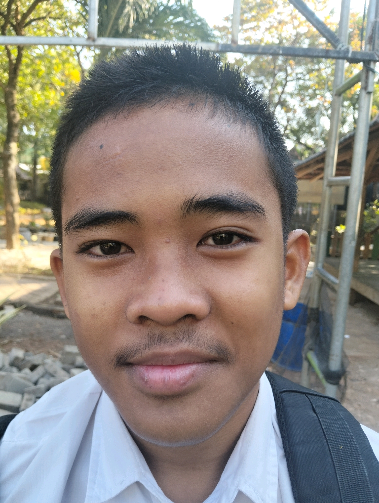 In an outdoor, sunlit environment in Paciran, Indonesia, a young East Asian male, appearing to be a student due to his white collared shirt and backpack, is captured in a close-up portrait. He has dark, short hair, a prominent forehead with a faint scar and a mole above his left eyebrow, and dark eyes with visible eyelashes. His eyebrows are thick and well-defined. A faint mustache is visible above his upper lip, which is slightly parted and appears dry. His skin tone is medium brown. Behind him, out of focus, are elements of the environment: trees with yellow and green foliage, suggesting a tropical setting, and metal structures that could be part of a building or scaffolding. The ground is partially visible and appears to be made up of dirt and possibly some construction debris. The lighting indicates it is daytime, with sunlight casting some shadows and highlights.