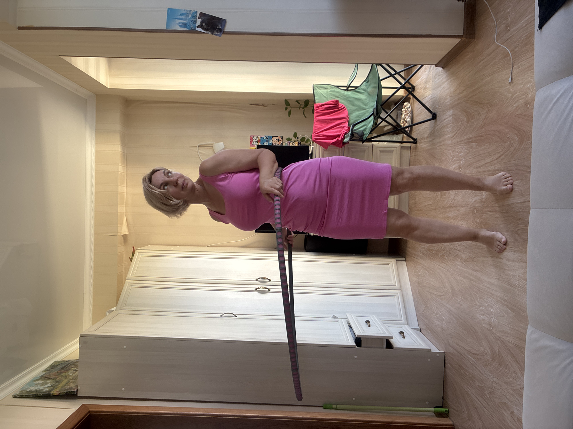 A Caucasian woman with blonde bob-cut hair stands in a room, holding a hula hoop horizontally with both hands. She is wearing a sleeveless pink dress. The room has light-colored walls with vertical stripes and a light-colored wooden floor. To the left, there is a tall, light-colored wardrobe with multiple doors and drawers. A small painting hangs on the wall above the wardrobe. To the right of the woman, there is a green folding chair with a pink t-shirt draped over it, and a small table with a plate of food. A television screen is visible behind the chair. The scene appears to be indoors, possibly an apartment in Sochi, Russia, judging by the context provided.