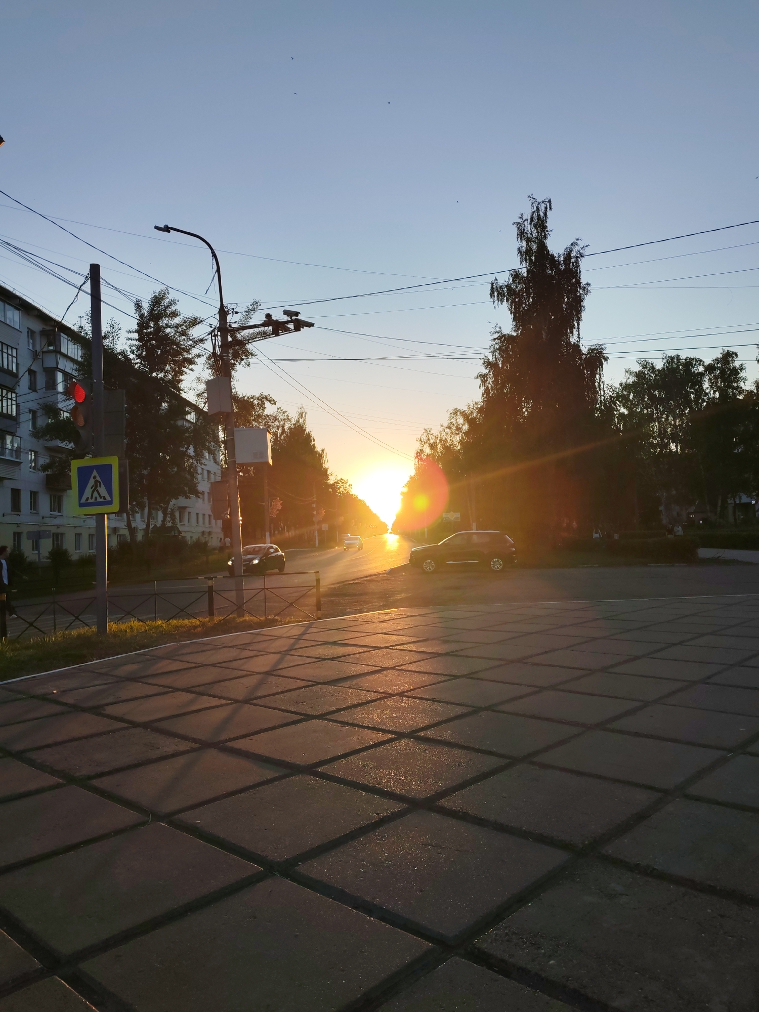 A photograph captures a street scene in the city of Chaikovsky, Russia, during sunset. The sun, a bright orb low on the horizon, casts long shadows and a warm, golden light across the pavement and the street. The sky is a clear, pale blue.
In the foreground, large, square paving stones form a sidewalk or pedestrian area, with a pattern of shadows cast by an unseen fence or railing. To the left, a traffic light is visible with a red signal illuminated, and a pedestrian crossing sign with a blue square and white pictogram. A multi-story building with several windows lines the street on the left.
A few cars are present on the road, one black car is parked or moving slowly on the right side of the road, and a white car is further down the street. Trees with dark foliage are silhouetted against the bright sky and the setting sun on the right side of the image. Overhead, numerous power lines crisscross the sky, supported by utility poles. A few small birds are visible in the sky. The overall impression is of an urban environment bathed in the warm glow of the setting sun.