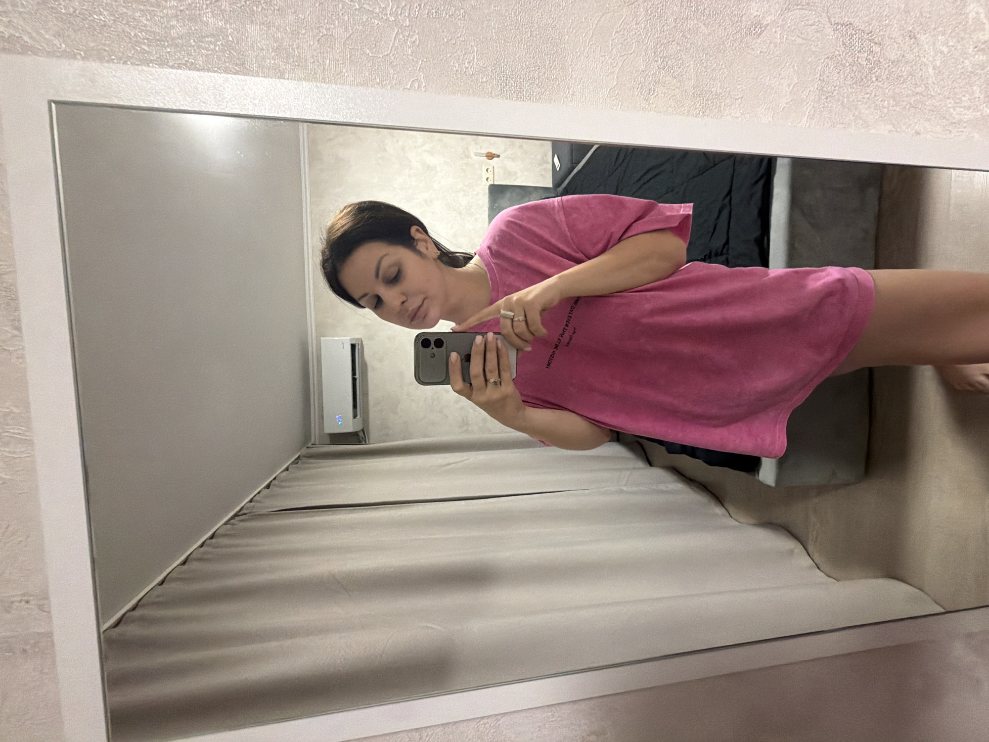A woman is taking a selfie in a mirror in what appears to be a bedroom. She is wearing a bright pink oversized t-shirt and has dark hair pulled back. She is holding her phone in her left hand and her right hand is resting on her chin. In the reflection, a bed with dark bedding and a grey headboard is visible. To the left, there are floor-to-ceiling beige curtains. An air conditioning unit is mounted on the wall above the curtains. The wall to the right of the mirror has a textured, light-colored wallpaper. The overall environment suggests a domestic setting. There is no indication of the location being in Sochi, Russia.