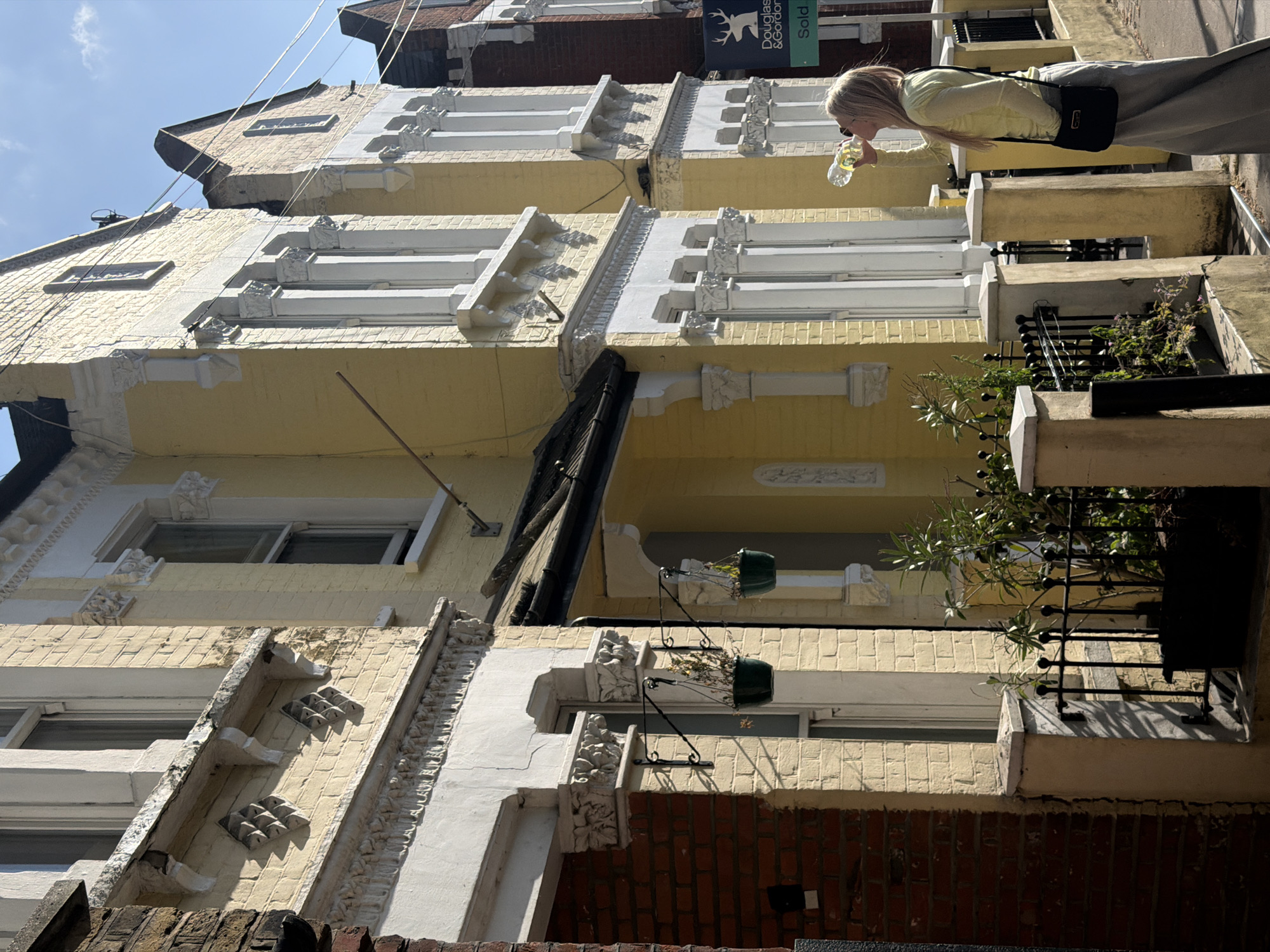 This rotated image captures a sunlit street scene, likely in London, United Kingdom, featuring a series of multi-story terraced buildings with a person in the foreground. The buildings are predominantly painted a pale yellow, contrasting with intricate white architectural details such as cornices, window frames, and decorative elements. Sections of exposed red brick are also visible, particularly at the lower levels and roofs.
In the mid-ground, a woman with blonde hair, dressed in a light yellow long-sleeved top, light-colored trousers, and carrying a black shoulder bag, is actively drinking from a clear plastic water bottle. She stands facing the buildings, with parts of black wrought-iron balconies and green potted plants visible in the foreground and on lower levels.
The bright daylight suggests a clear and sunny day, with strong shadows cast across the facades. A patch of blue sky with some white clouds is visible at the top left. Notable details include stylized decorative patterns on the building's exterior and dark cables running across the upper part of the frame. On one of the upper floors, an estate agent sign is prominently displayed, featuring the text "Douglas & Gordon" with a stag logo, and "Sold" beneath it, which is characteristic of the London property market. The architecture, combined with the estate agent branding, strongly indicates the location context of London.