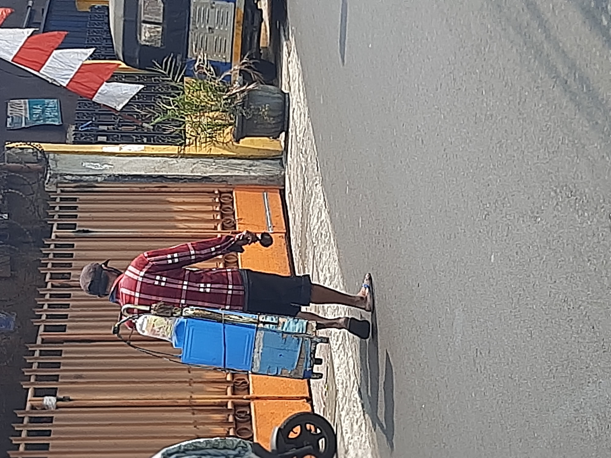 An Indonesian man wearing a red and white plaid shirt, dark shorts, a dark cap, and flip-flops walks along a paved road in Central Jakarta, Indonesia. He carries a blue cooler on his left shoulder, supported by straps, and holds a brown spherical object in his right hand. The man is walking away from the camera. Behind him, a closed orange metal gate stands on the left side of the image, and a building with red and white flags is visible on the right. A small motorized vehicle is parked to the right of the flags. The road has a textured asphalt surface, and a faint shadow of the man and his load stretches across it. The scene appears to be set in a commercial or residential area, with buildings and fencing lining the street.