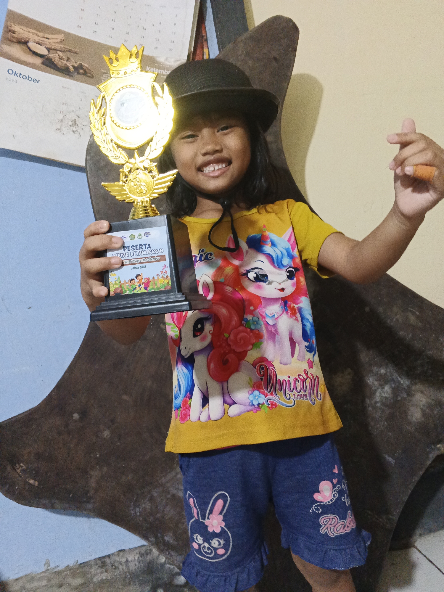 A young Indonesian girl wearing a yellow t-shirt with unicorns and blue shorts with rabbits is smiling and holding up a trophy. She is also wearing a black hat and is making a heart shape with her fingers. The trophy has a gold crown and a blue plaque with text that appears to be in Indonesian. In the background, there is a calendar showing October 2023, and a large, dark, textured object that resembles a piece of furniture or a sculpture. The overall scene suggests a celebratory moment, possibly after a competition or event. The location is identified as Warungkiara, Indonesia.